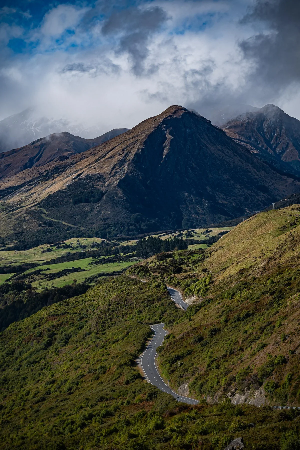 A winding mountain road through green hills leading toward tall, rugged mountains under a partly cloudy sky.