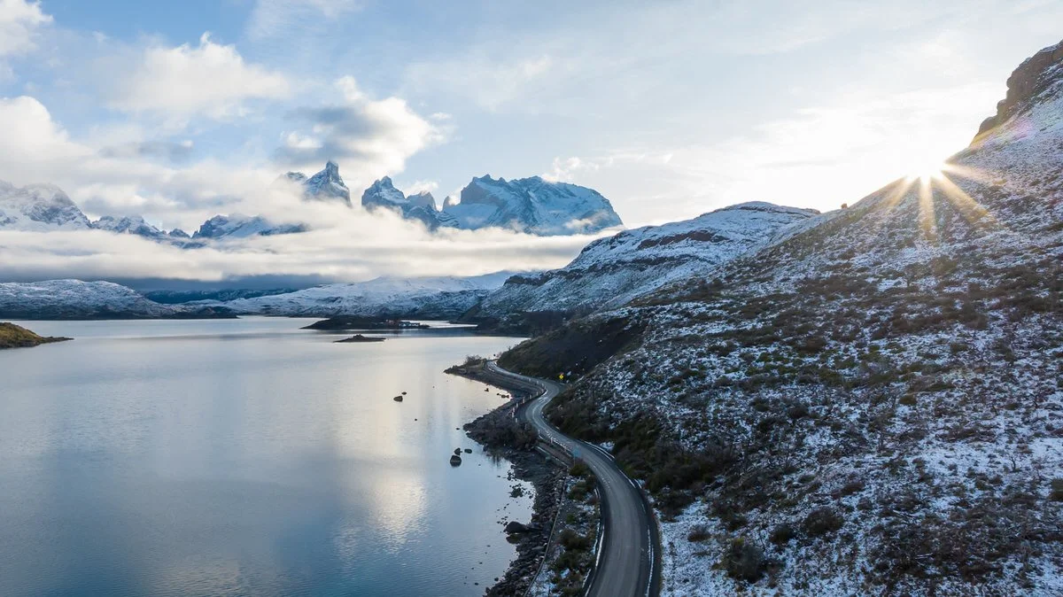 A winding road along a lake with snow-covered hills and mountains in the background, with the sun shining over the horizon.