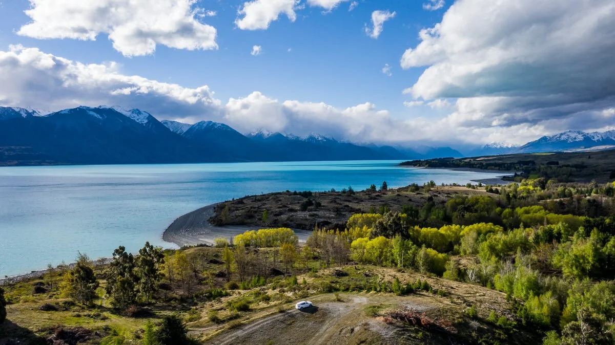 Scenic view of a large lake with snow-capped mountains in the background, surrounded by green trees and hills under a partly cloudy sky.