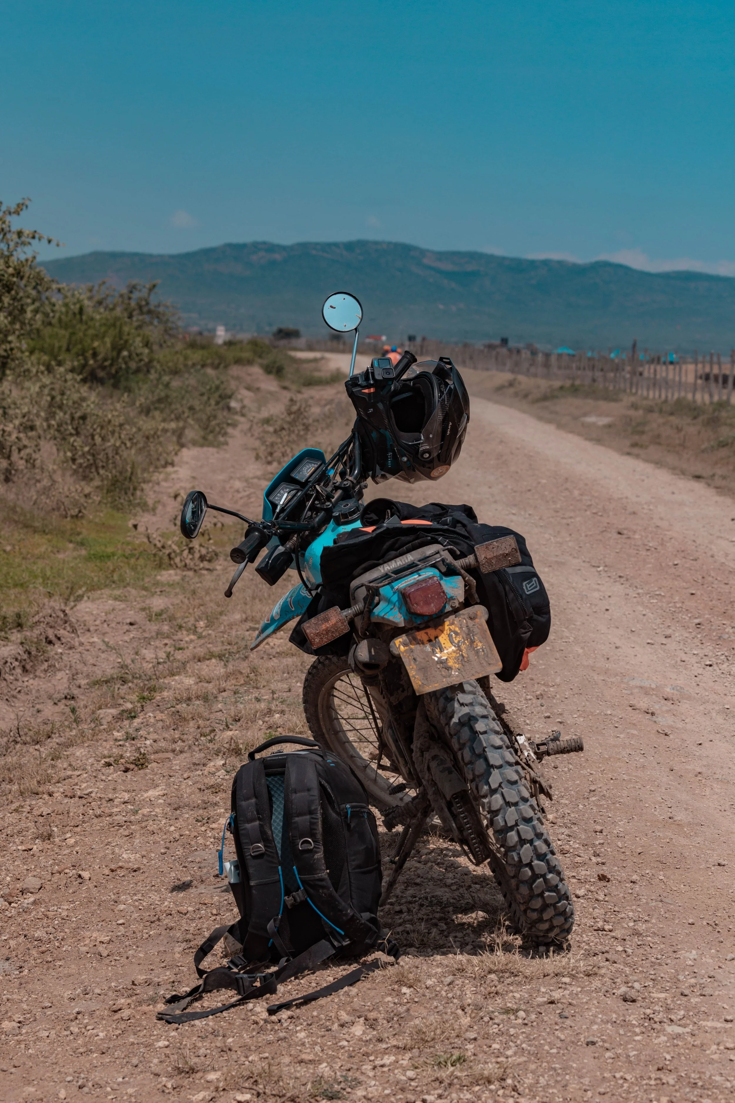 A dirt bike with a backpack and saddle bags parked on a remote dirt road, with mountains in the background under a clear blue sky.