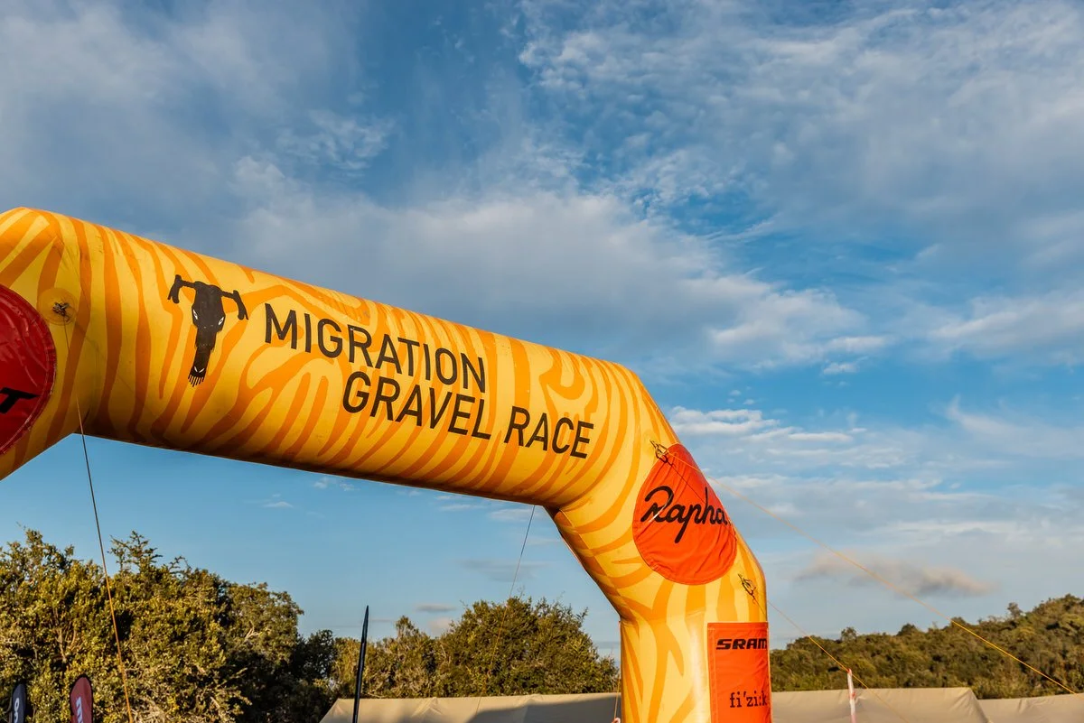 Yellow inflatable arch with orange zebra pattern, labeled 'Migration Gravel Race' and sponsored by Rapha, trimodated over a landscape of trees and a partly cloudy sky.