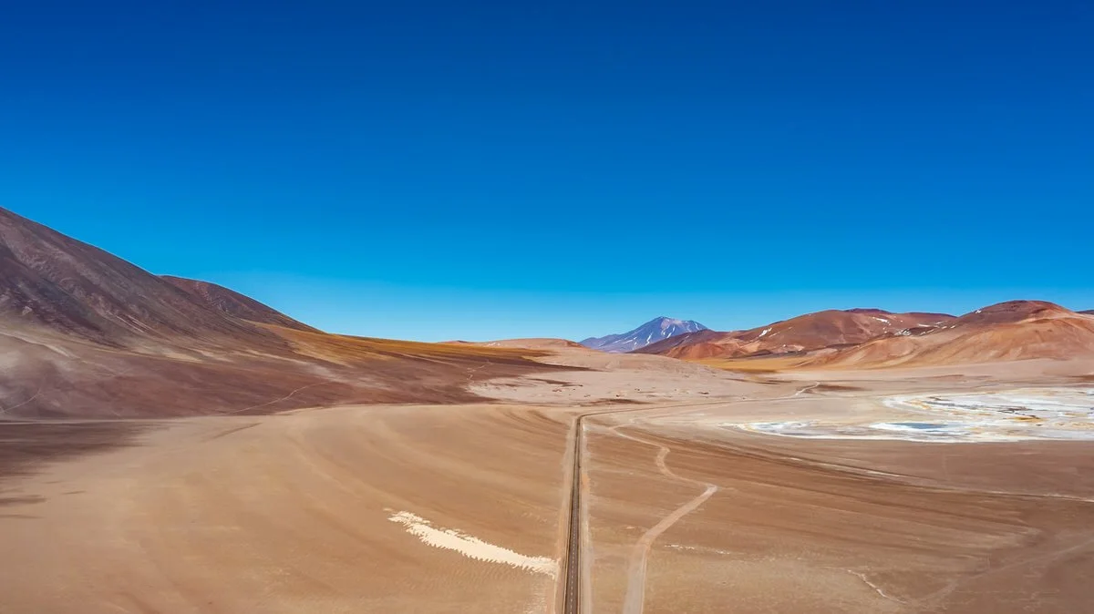 A vast desert landscape with distant mountain ranges under a clear blue sky, featuring dry, sandy terrain and sparse vegetation.