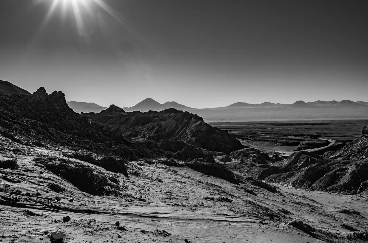 A black and white photograph of a desert landscape with rugged mountains, a wide flat plain, and a bright sun in the sky.