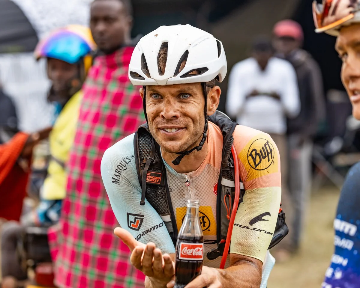 A male cyclist wearing a white helmet and colorful cycling gear holding a Coca-Cola bottle, smiling.