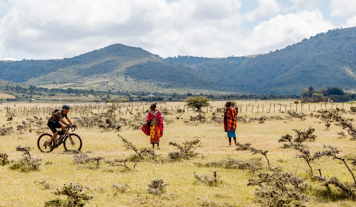 A person riding a bicycle past two women dressed in colorful traditional Maasai clothing in an open grassland with mountains in the background.