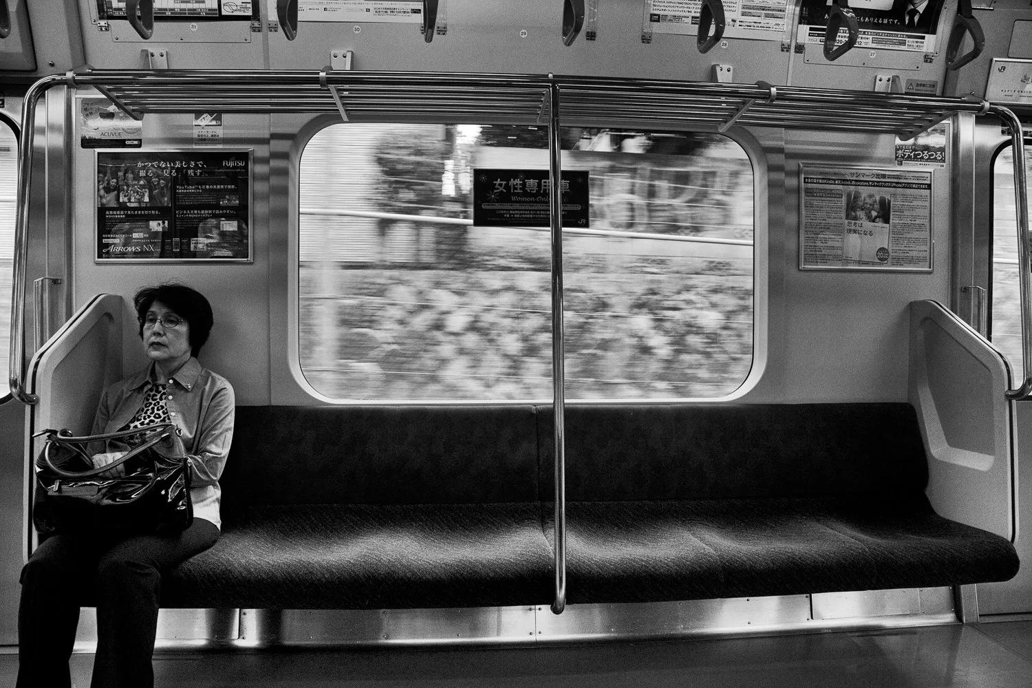 A woman sitting alone on a train or subway with a bag on her lap, looking away from the camera, with an empty seat next to her, and blurred scenery outside the window.