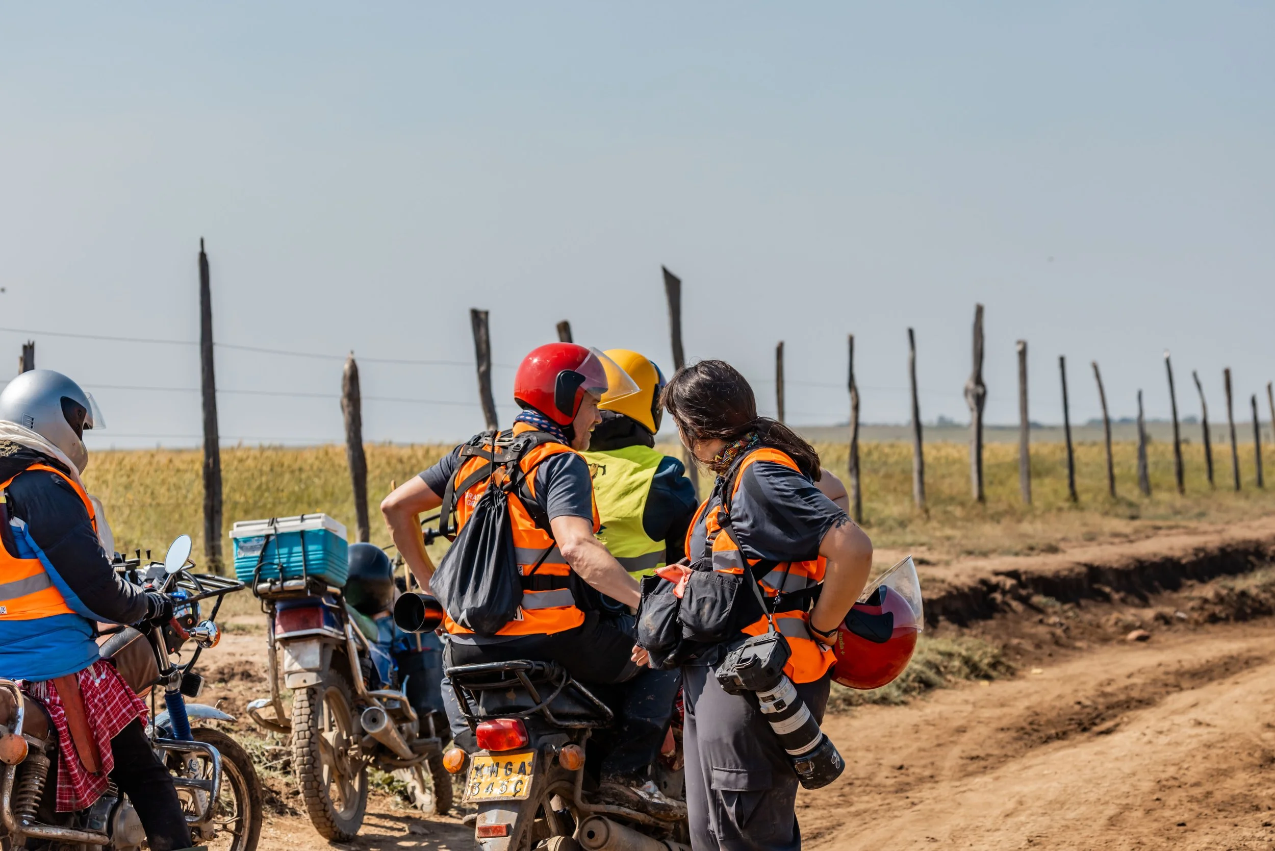 Group of people in safety gear and helmets gathered around a motorcycle on a dirt road, with a rural landscape and wooden fence in the background.