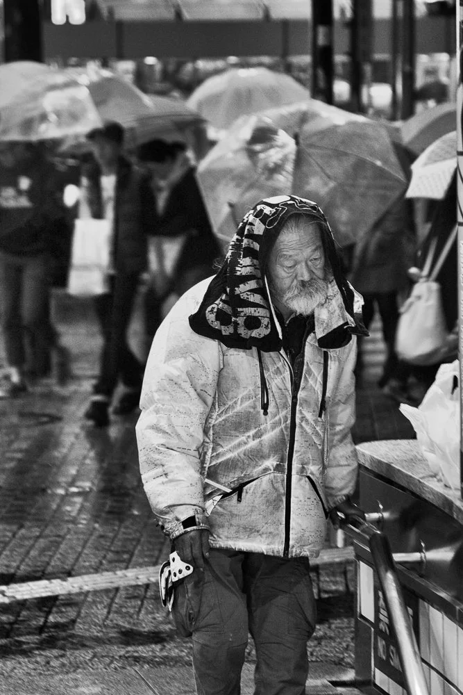 An older man with a beard and mustache wearing a jacket with a hood over his head, walking in the rain at night with several people and umbrellas in the background.