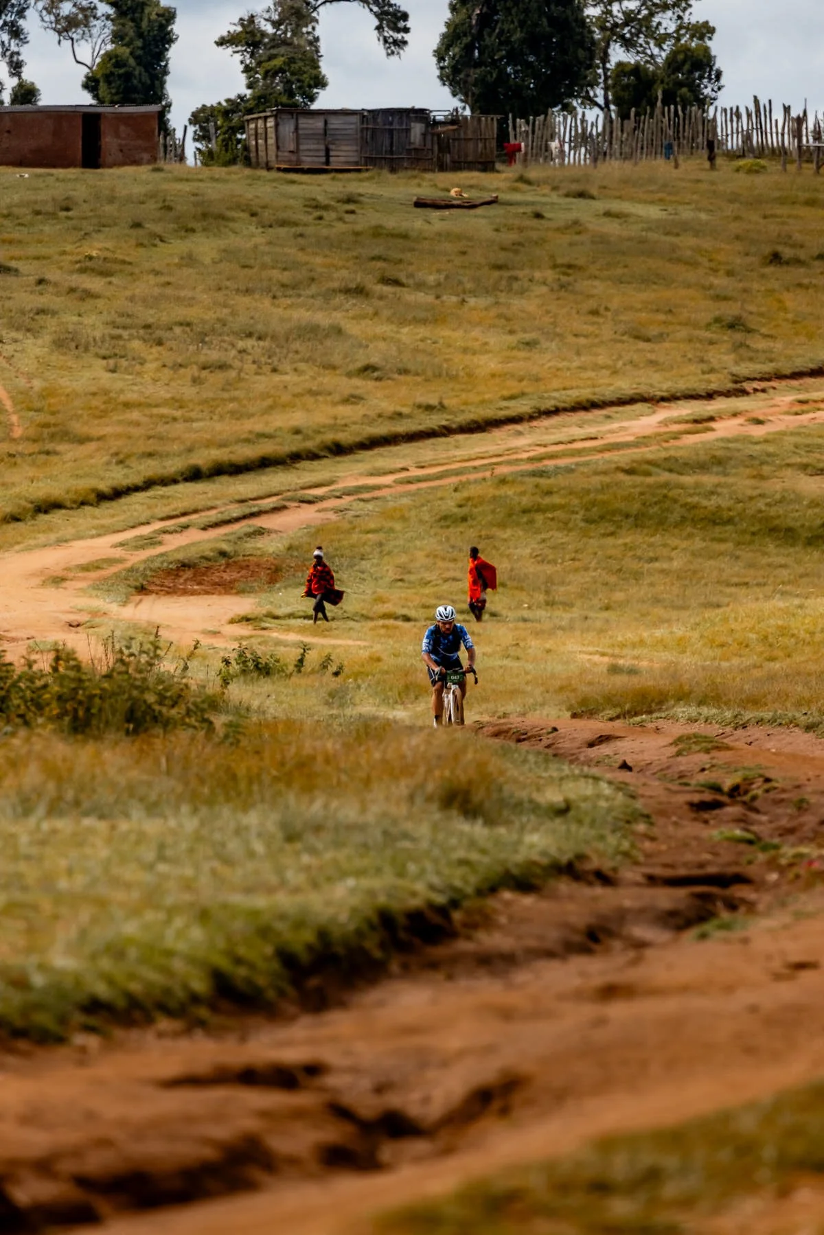 A cyclist riding on a dirt trail in a grassy, hilly landscape with people walking in the background and small houses with wooden fences at the top of the hill.