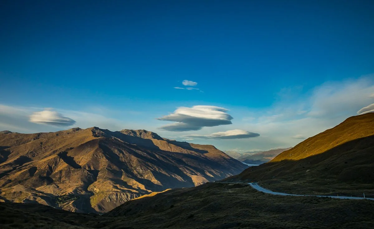Scenic view of a mountain valley with a winding road, hills, and mountains under a blue sky with scattered clouds.