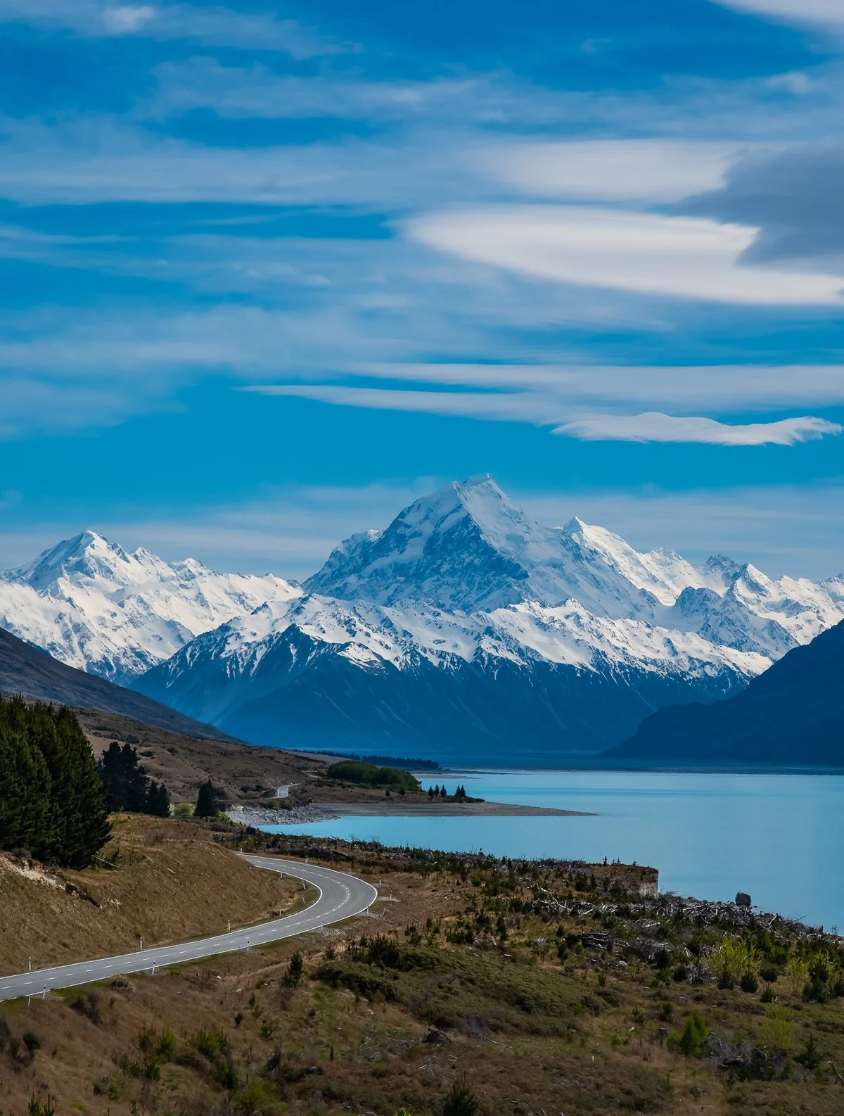 Snow-capped mountains near a body of water with a winding road in the foreground under a partly cloudy sky.