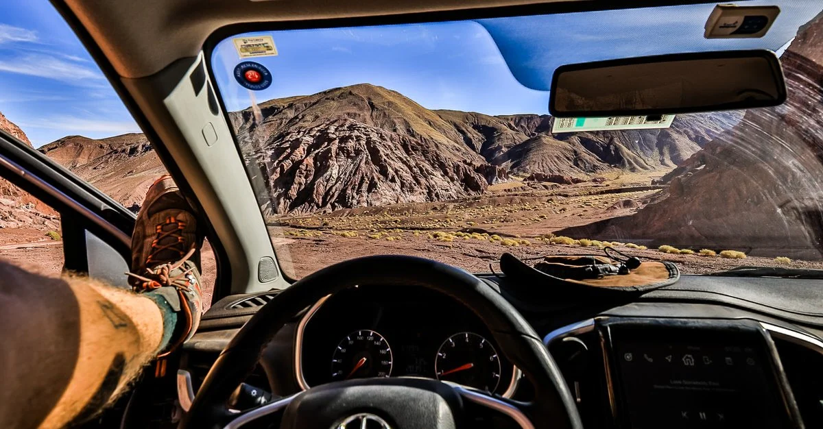 Inside a vehicle driving through a mountainous desert landscape with bare rocky hills under a clear blue sky.