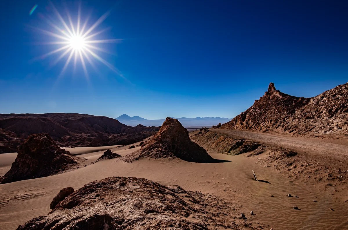 A bright sun shining over a desert landscape with sandy dunes and rocky formations under a clear blue sky.