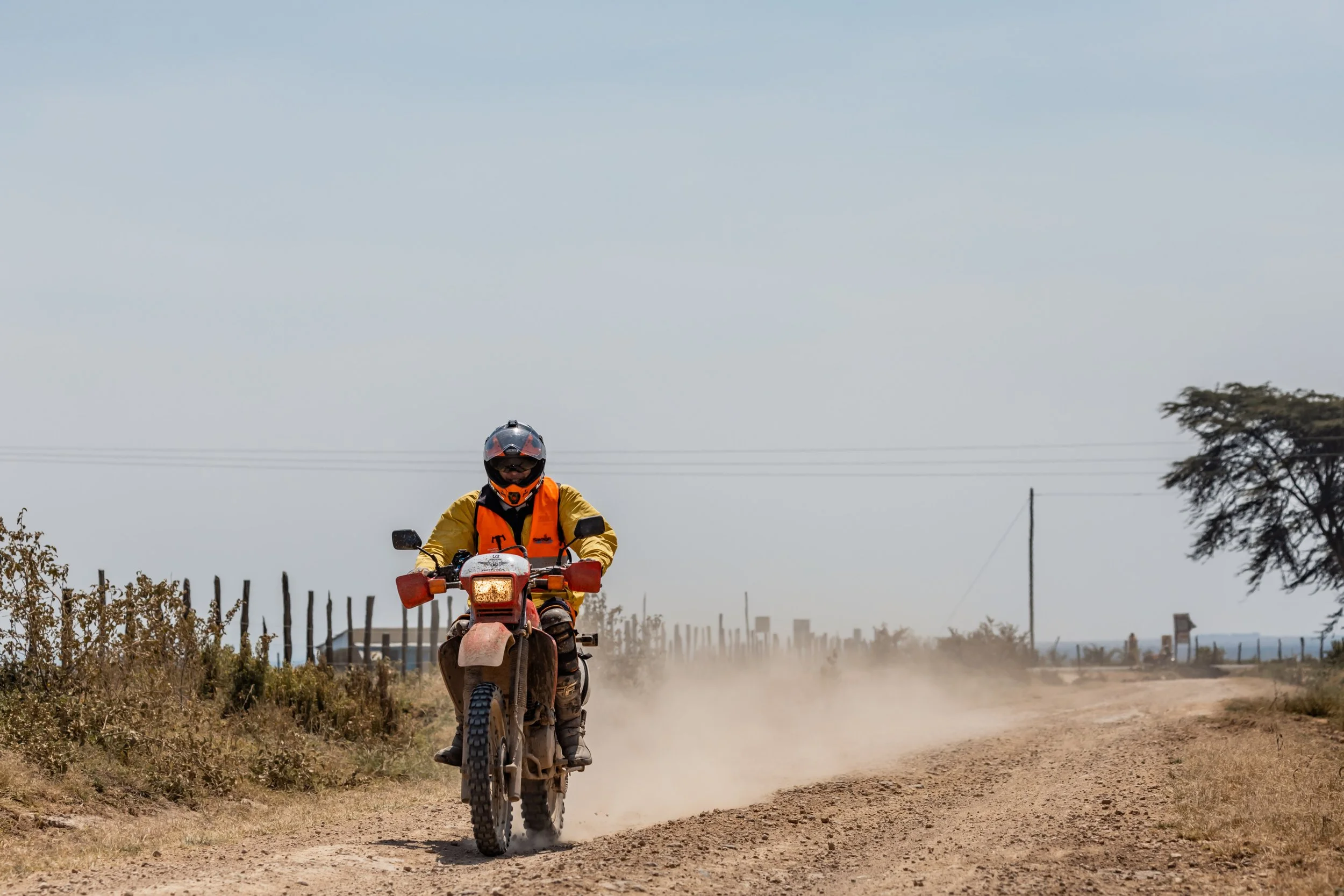 A person riding a dirt bike on a dusty rural road with fencing and sparse vegetation on both sides under a clear sky.