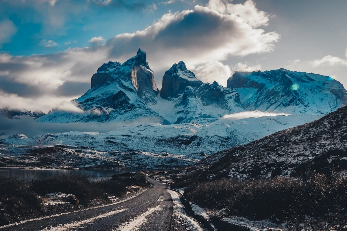 Snow-capped mountains in the distance with a dirt road leading towards them, under a cloudy sky.
