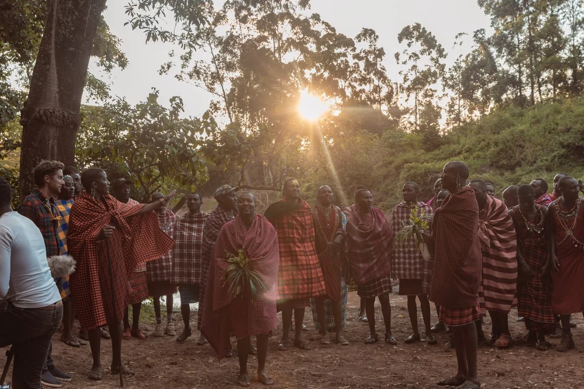 A group of Maasai people in traditional attire gathered outdoors during sunset, engaged in a communal event amidst trees and greenery.
