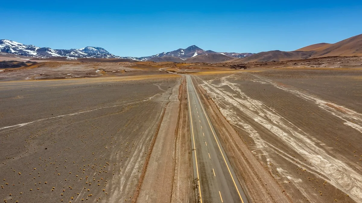 An empty road stretches through a barren desert landscape with mountains in the background, some snow on their peaks, and a clear blue sky above.