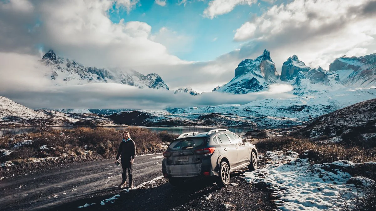 A person standing on a snowy roadside with rugged mountains and a lake in the background.