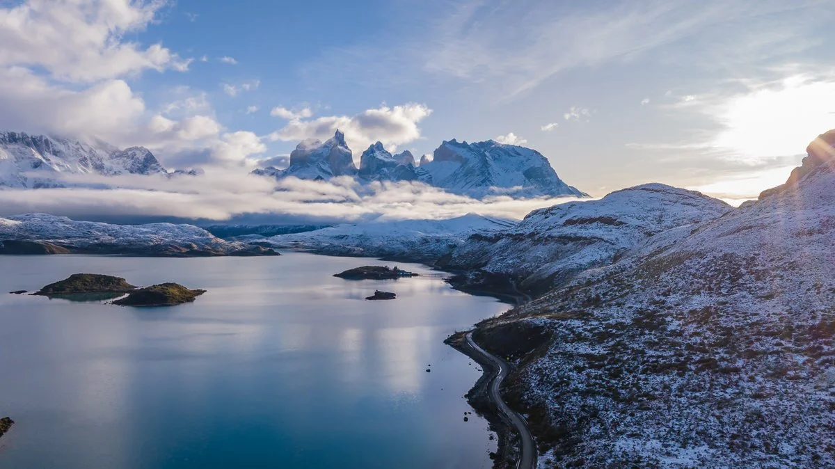Snow-capped mountains behind a lake with small islands and a winding road on the hillside