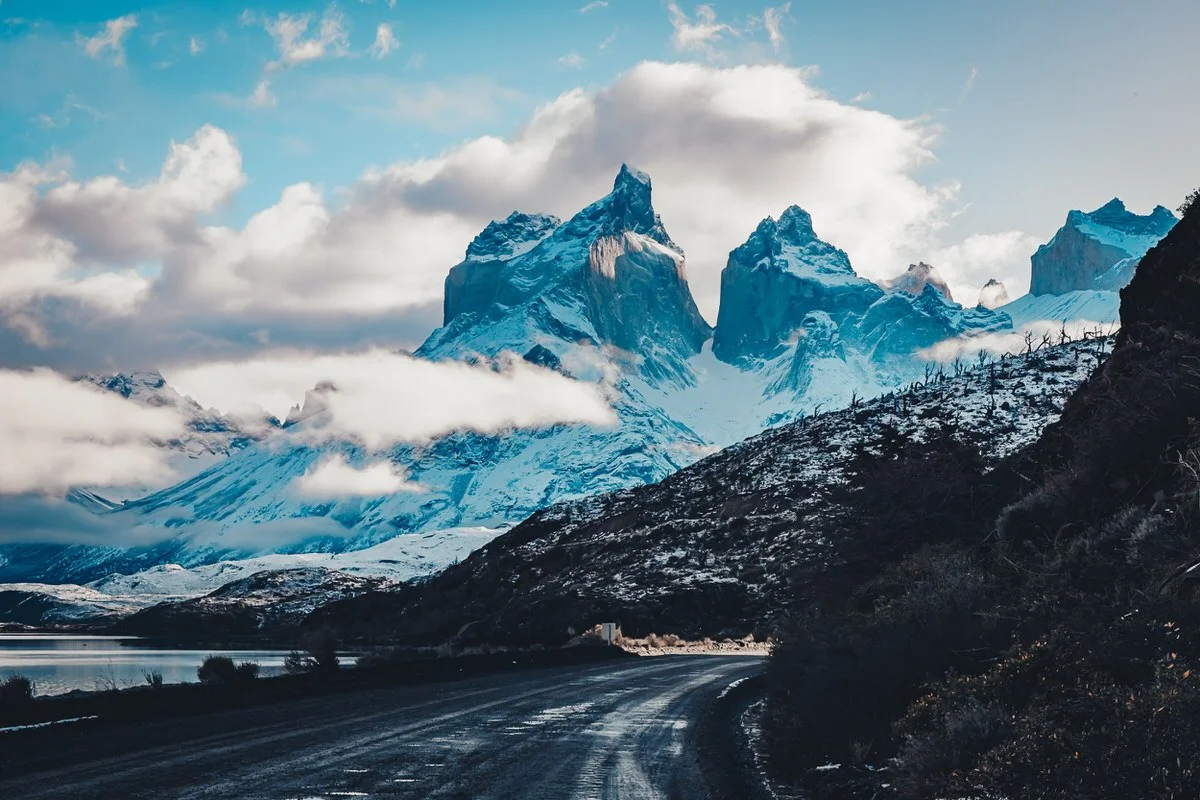 Snow-capped mountains in the distance with a winding gravel road in the foreground and some clouds in the sky.