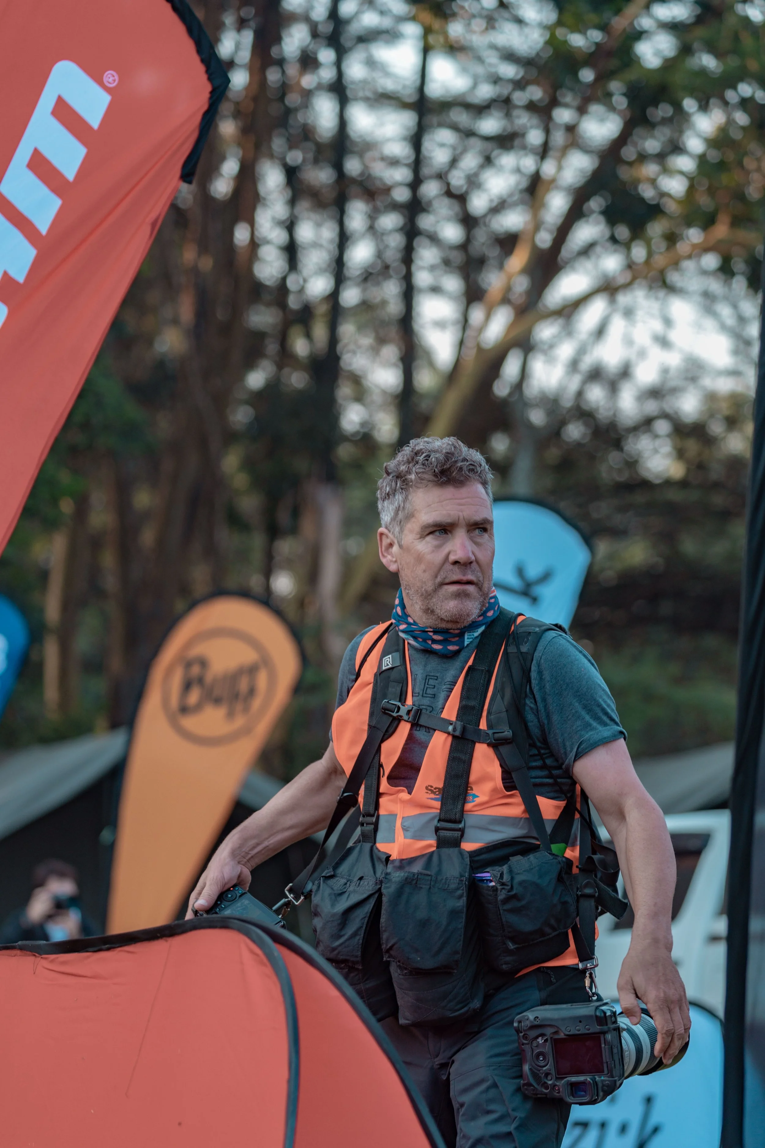 A man with short, curly gray hair and a beard wearing a backpack and holding a camera, standing outdoors at a sporting event or race, surrounded by orange and blue flags and trees in the background.