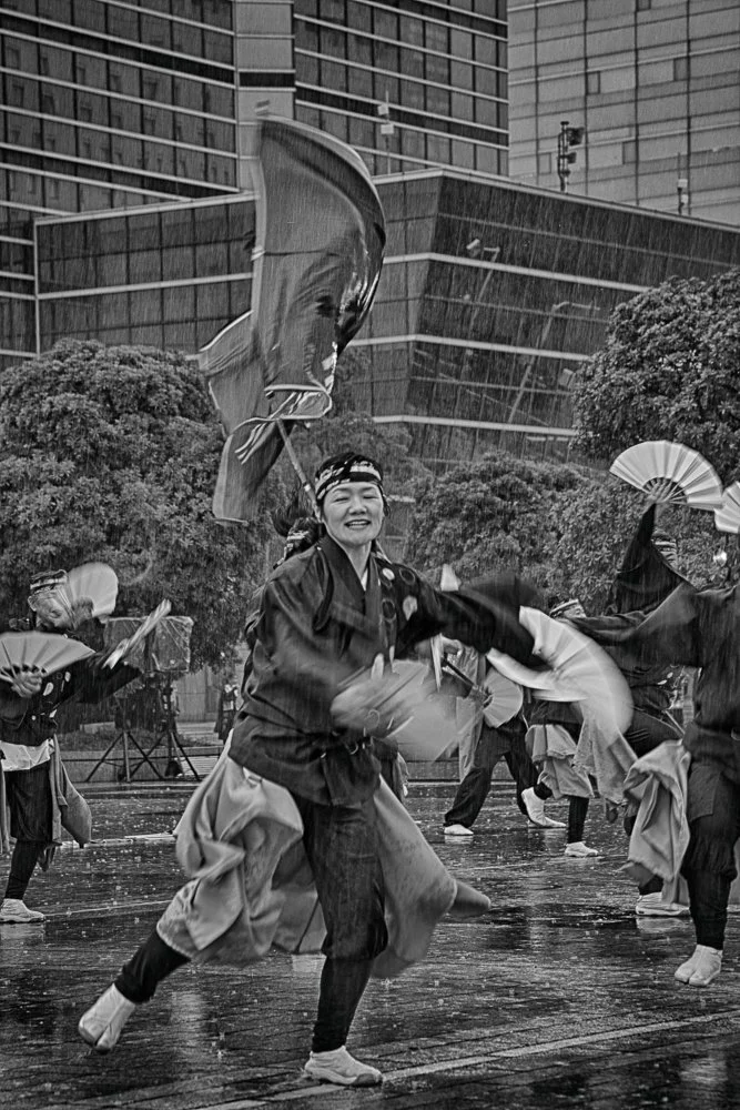 People dancing outdoors during a rainstorm, holding umbrellas and fans, with modern high-rise buildings in the background