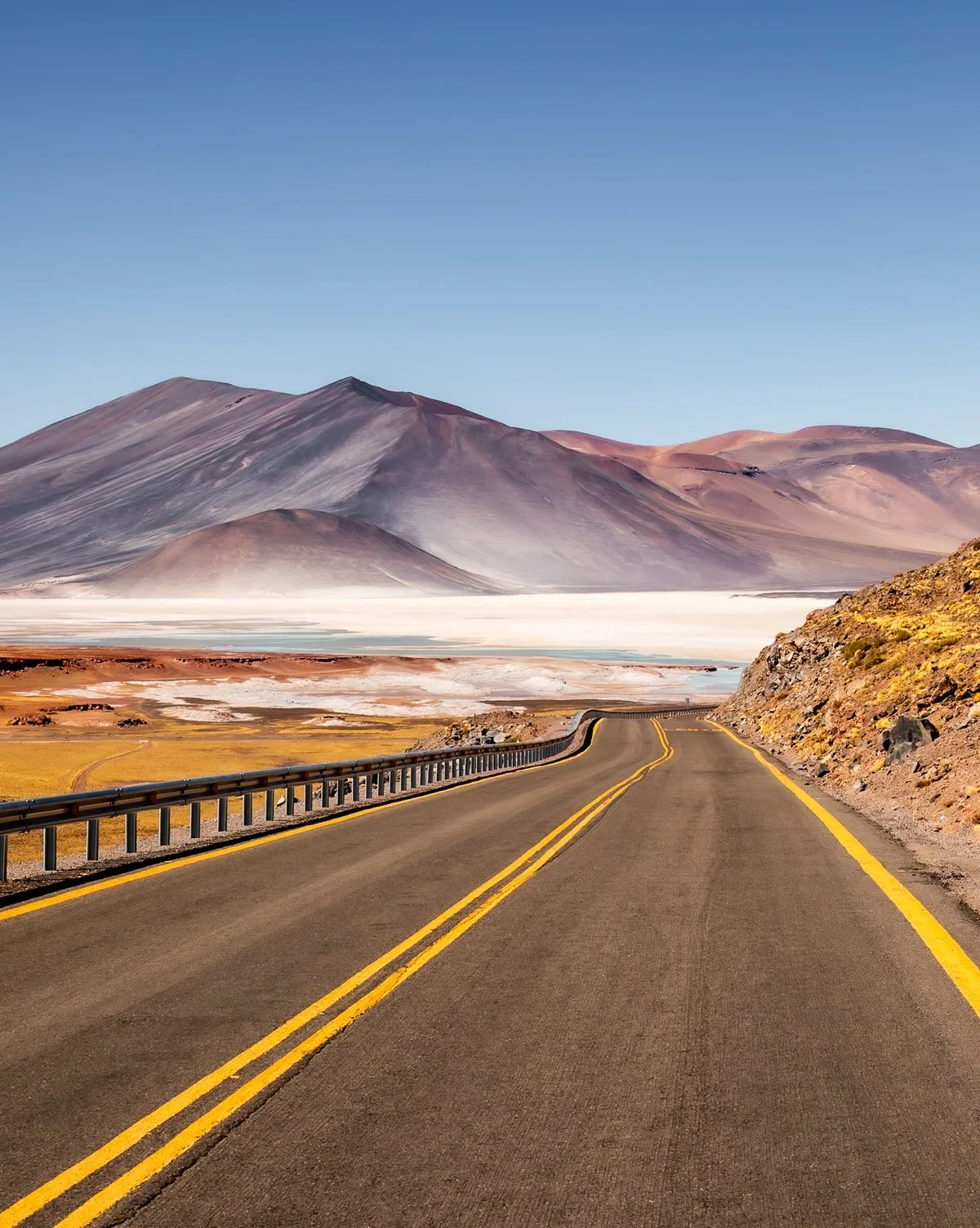 A scenic mountain road with a double yellow line travels through a desert landscape with colorful mountains in the background under a clear blue sky.