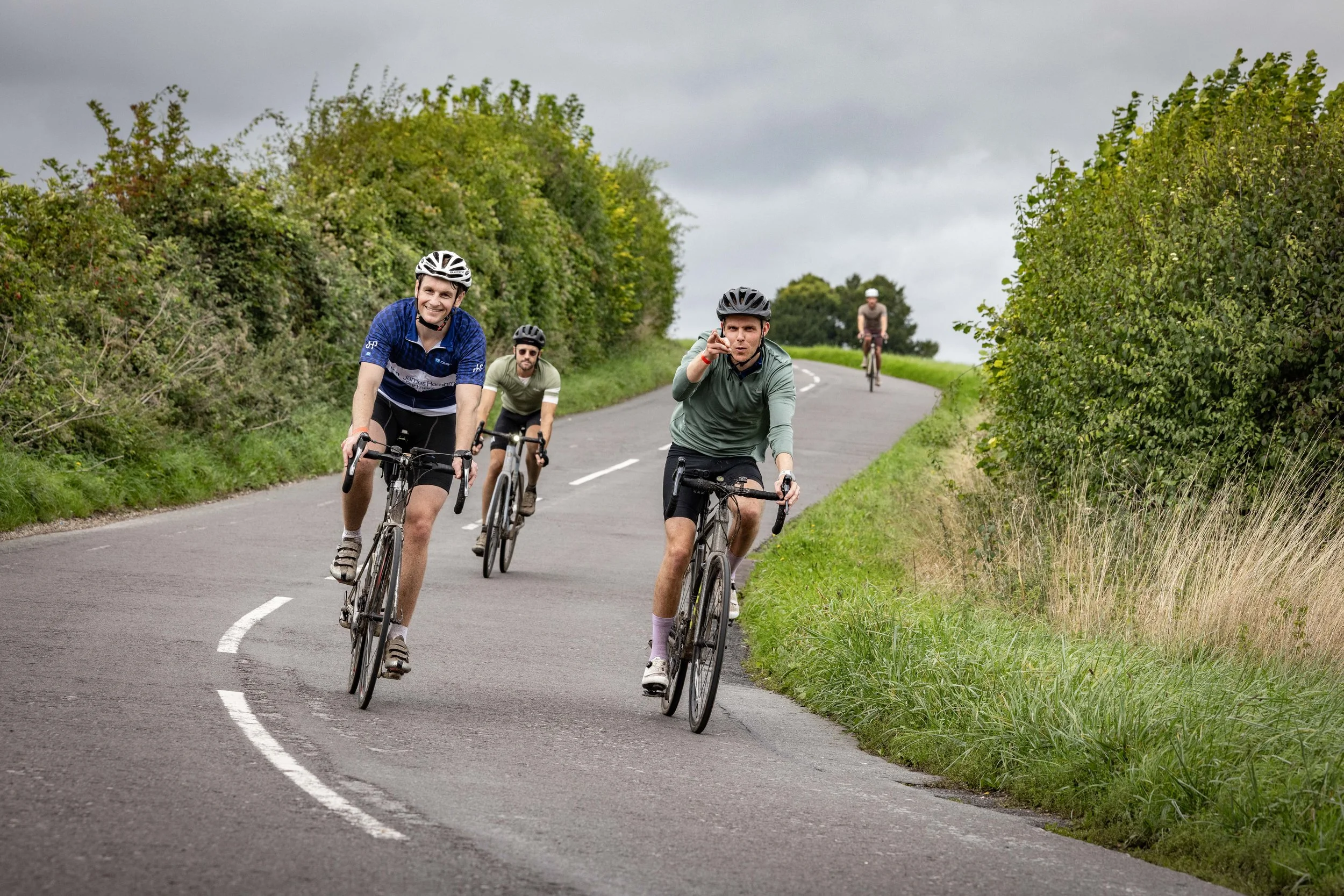 Four men riding bicycles on a winding countryside road with green bushes and cloudy sky.