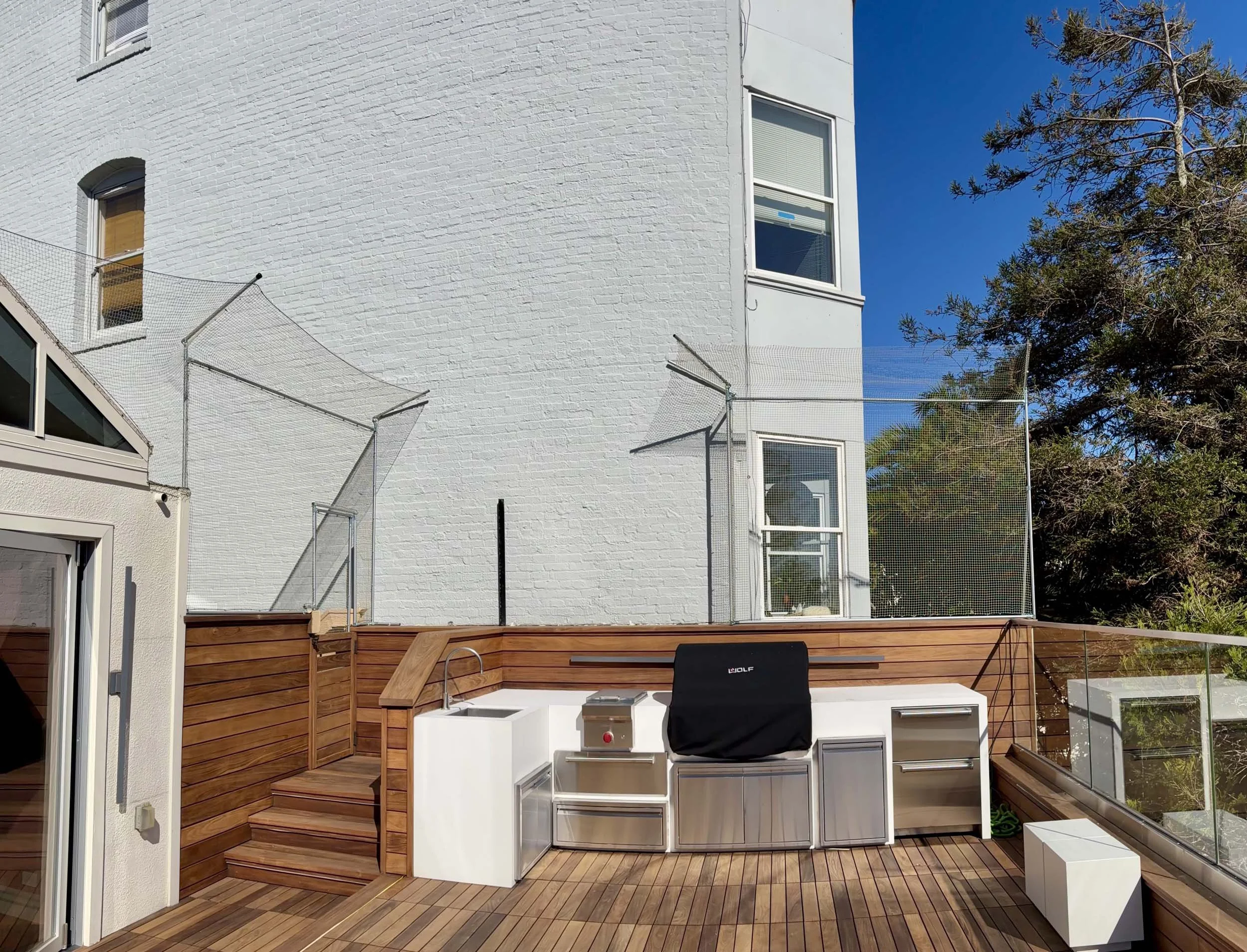 A rooftop deck with a wooden floor and a built-in grilling area. The deck is enclosed with glass railings, and there's a white brick building in the background with a window. There are trees on the right side under a blue sky.