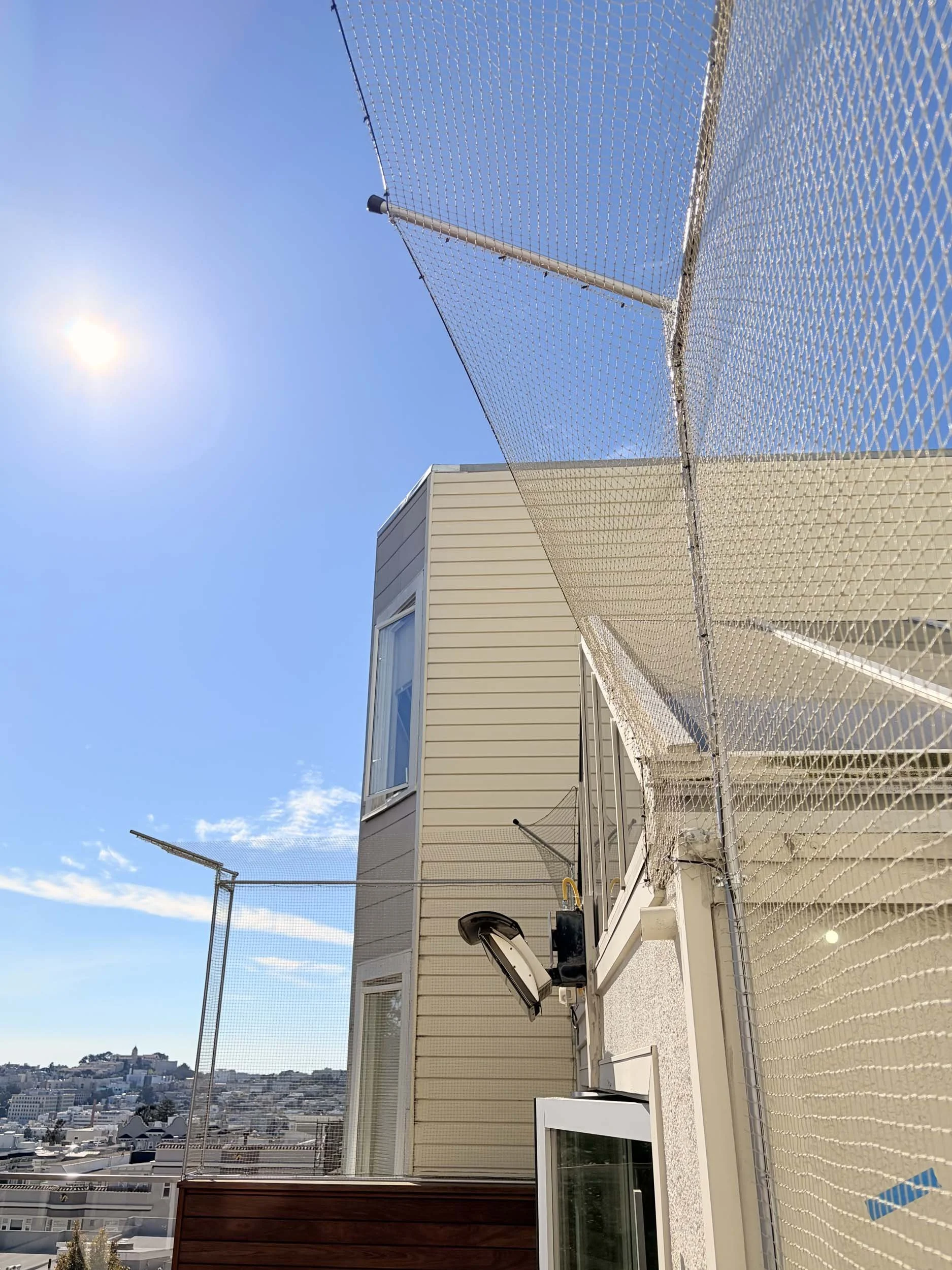 Apartment balcony with catio netting, a floodlight, and a cityscape in the distance under a clear blue sky with the sun.