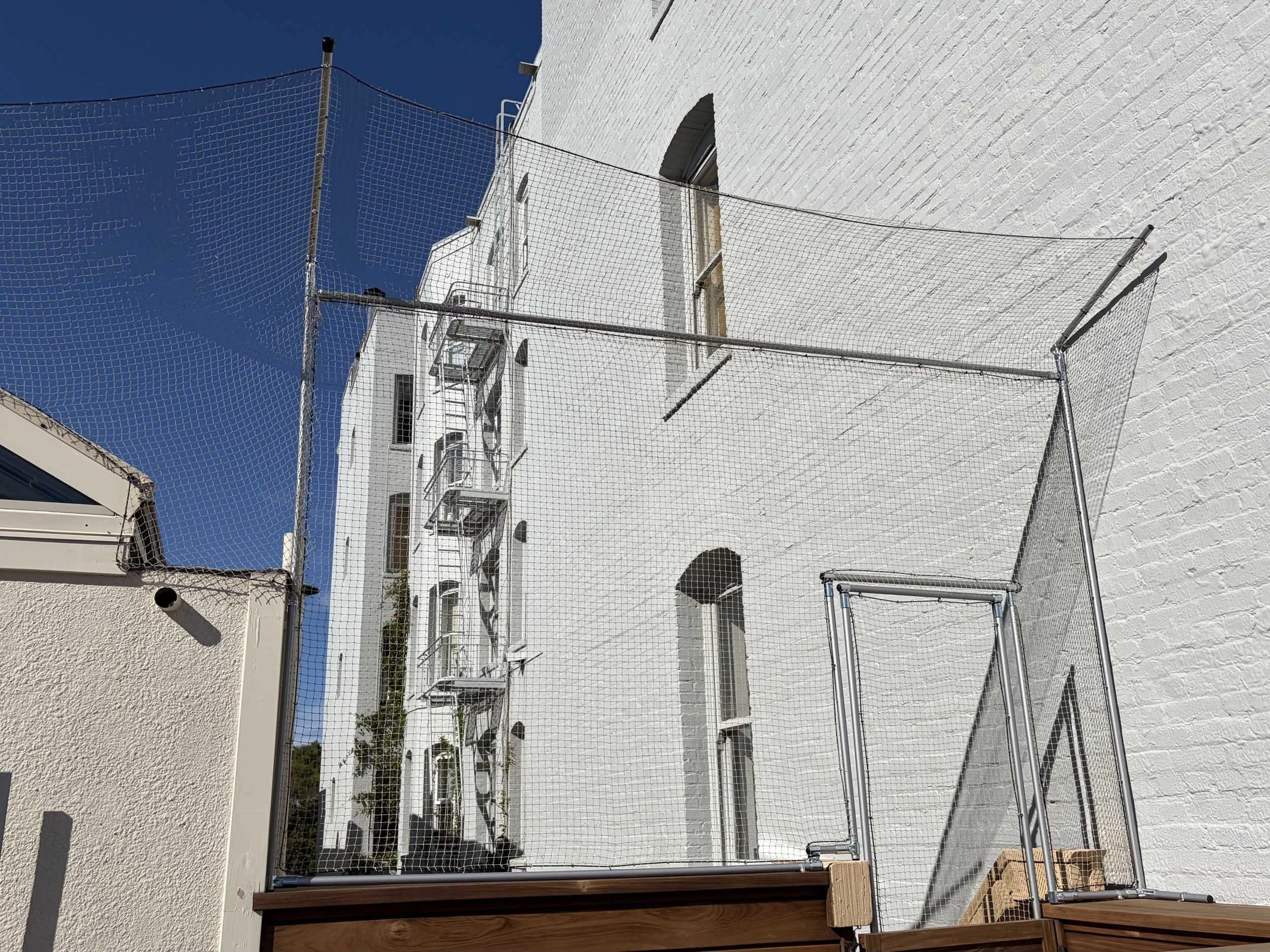 A white brick building with black arched windows and exterior fire escapes. There is a blue safety net hung on metal poles near the building, with clear blue sky in the background.