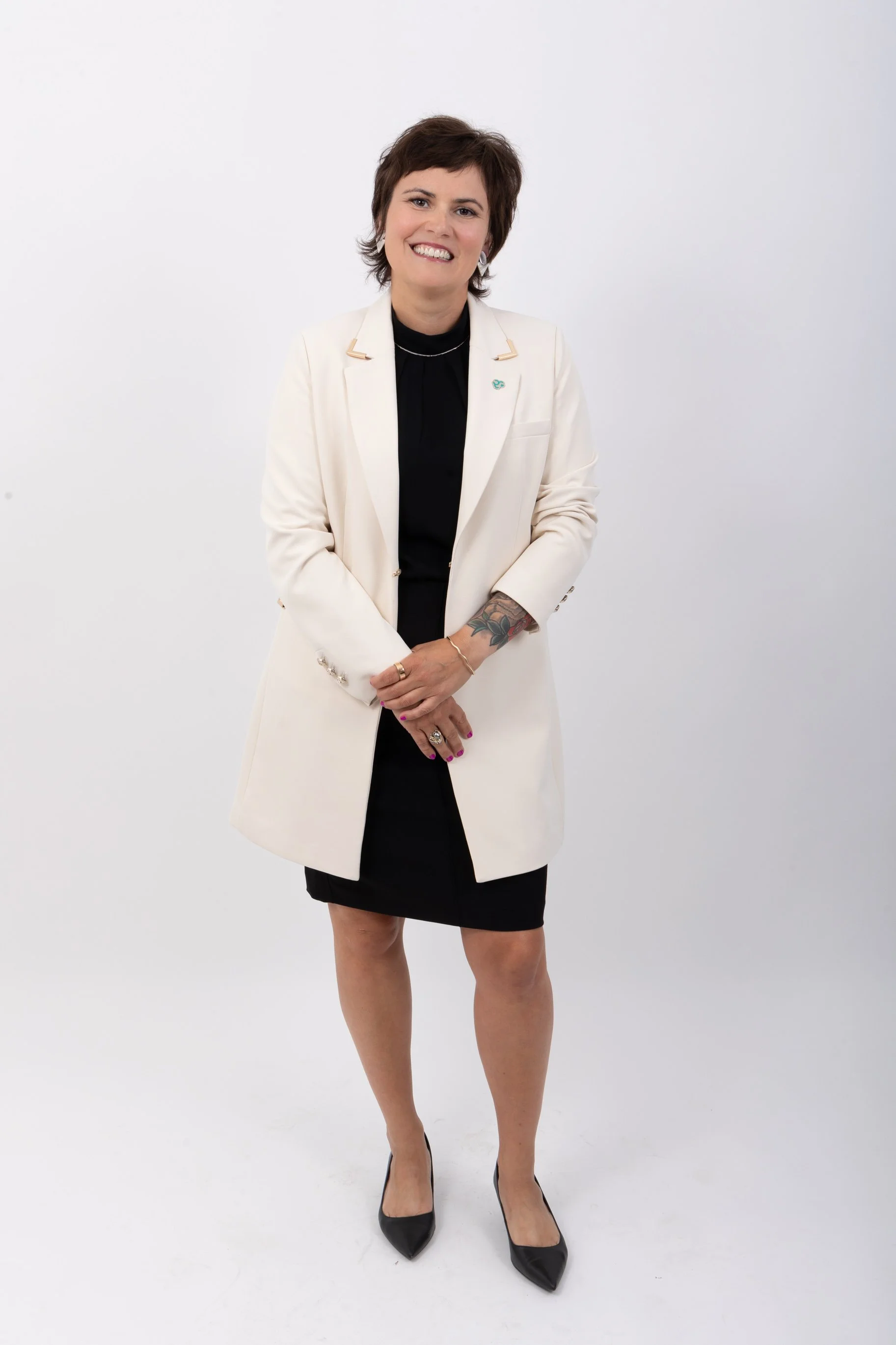 Femme souriante portant un tailleur blanc, une robe noire, des talons noirs, avec des tatouages visibles sur son bras et des bijoux.