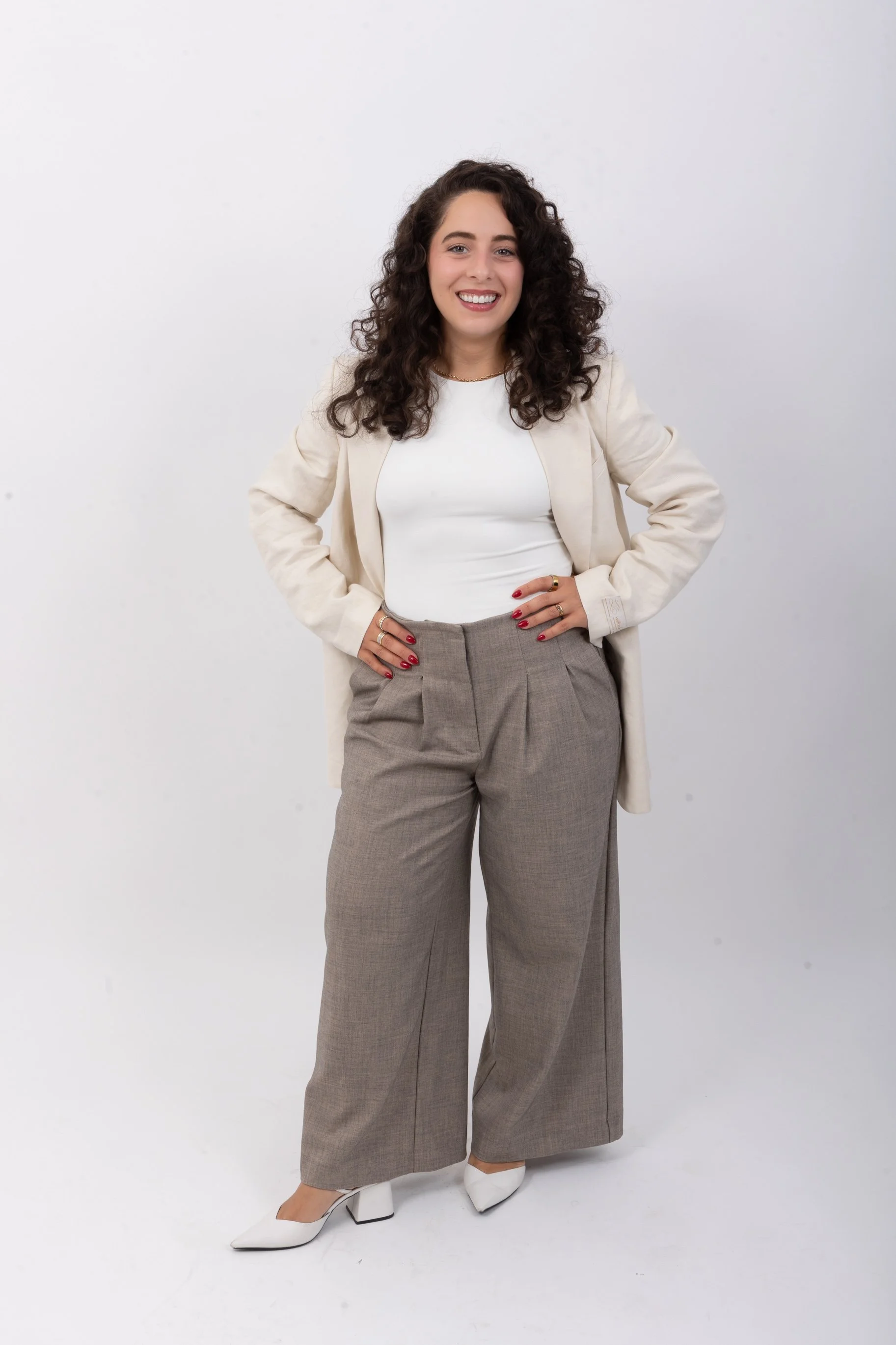 Une femme souriante avec des cheveux bouclés longs, portant une veste beige, un haut blanc, un pantalon gris large, et des chaussures blanches, posant devant un fond blanc.