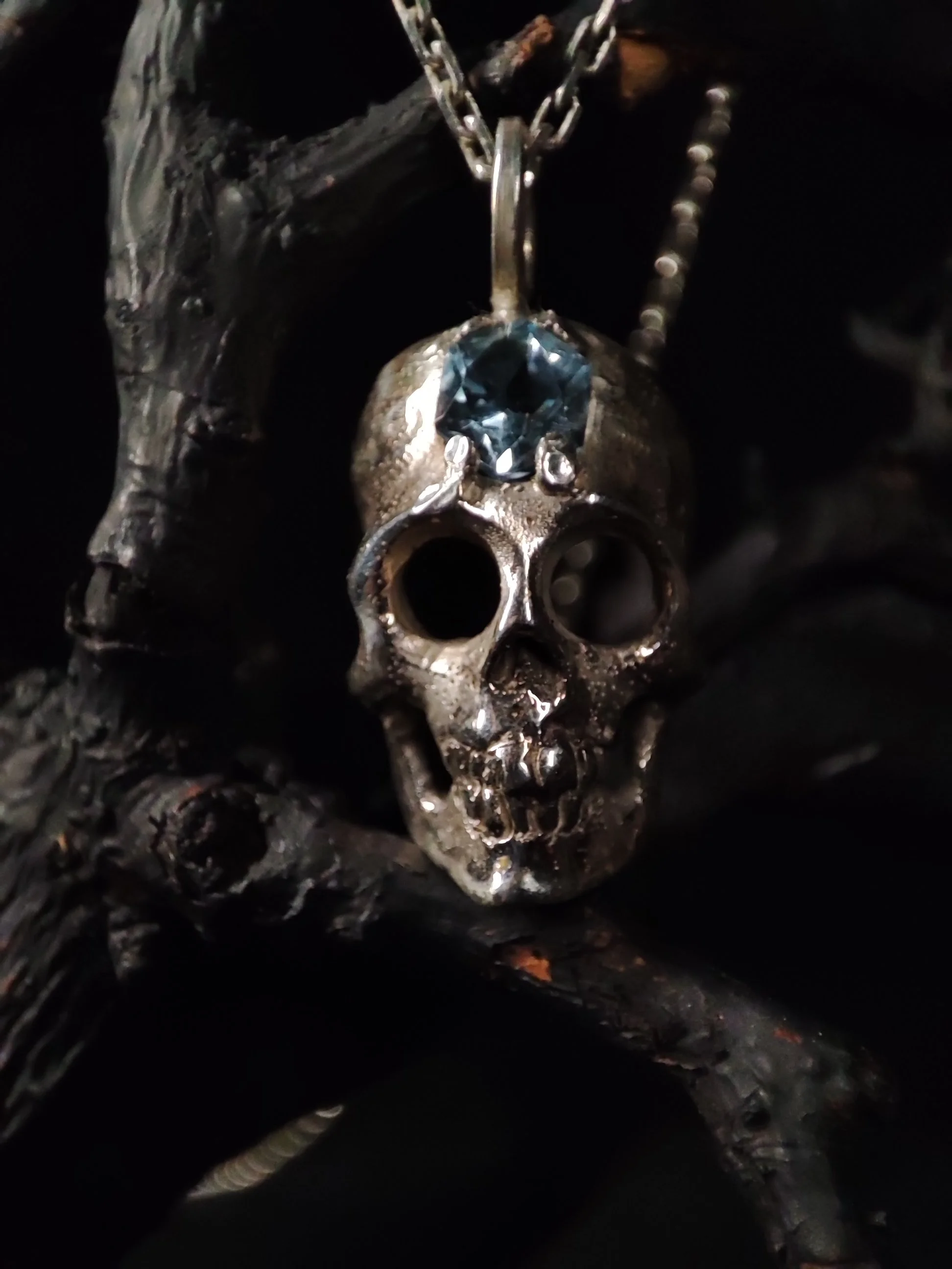 Un pendentif en forme de crâne en argent massif 925 création originale WIT joaillerie avec une pierre précieuse bleue, aigue marine, suspendu à une chaîne, posé sur du bois noir. Inspiré des saints des catacombes, Paris. 