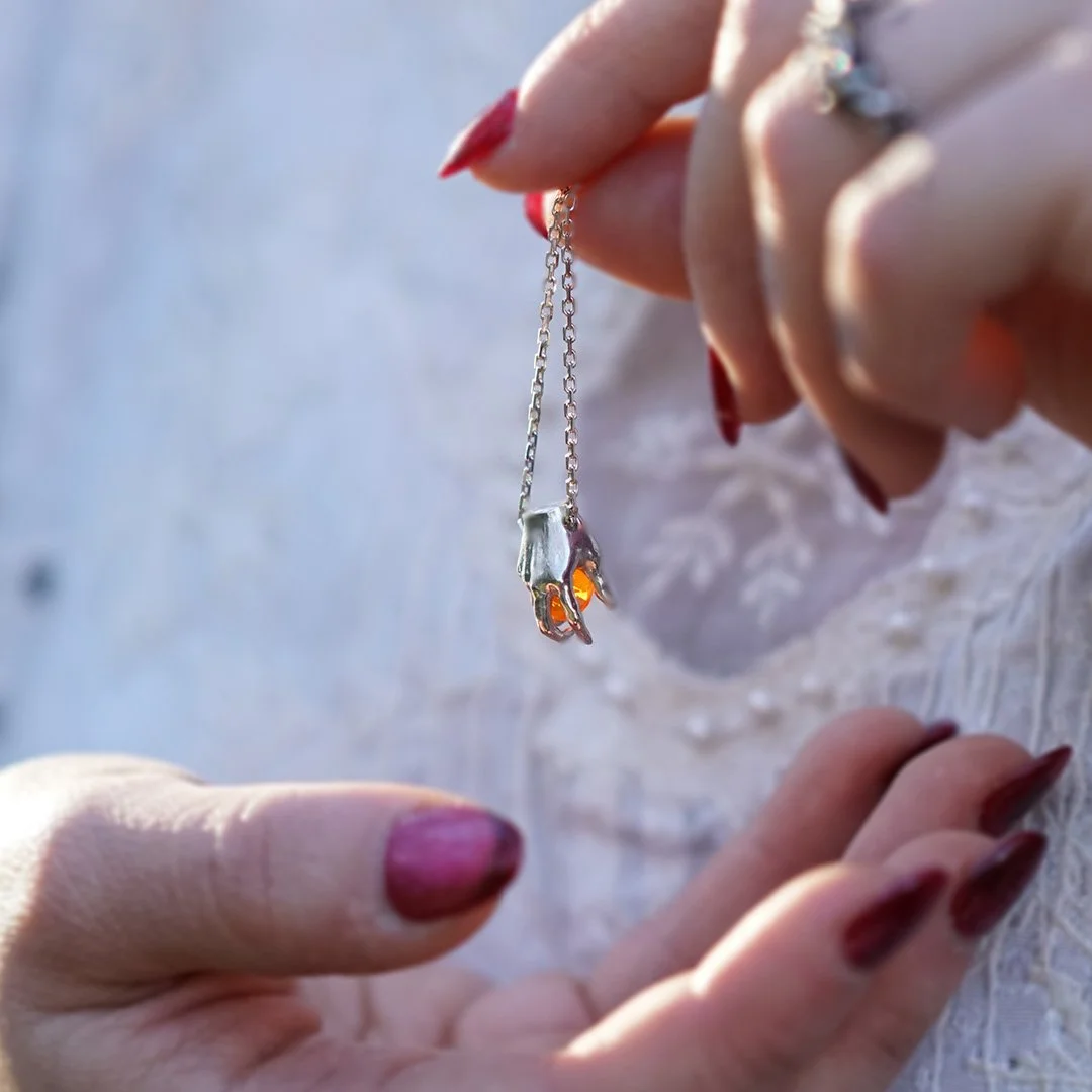Une main tenant un collier avec un pendentif en forme de main en argent massif 925 ornée d'une opale de feu. Création originale de WIT joaillerie.