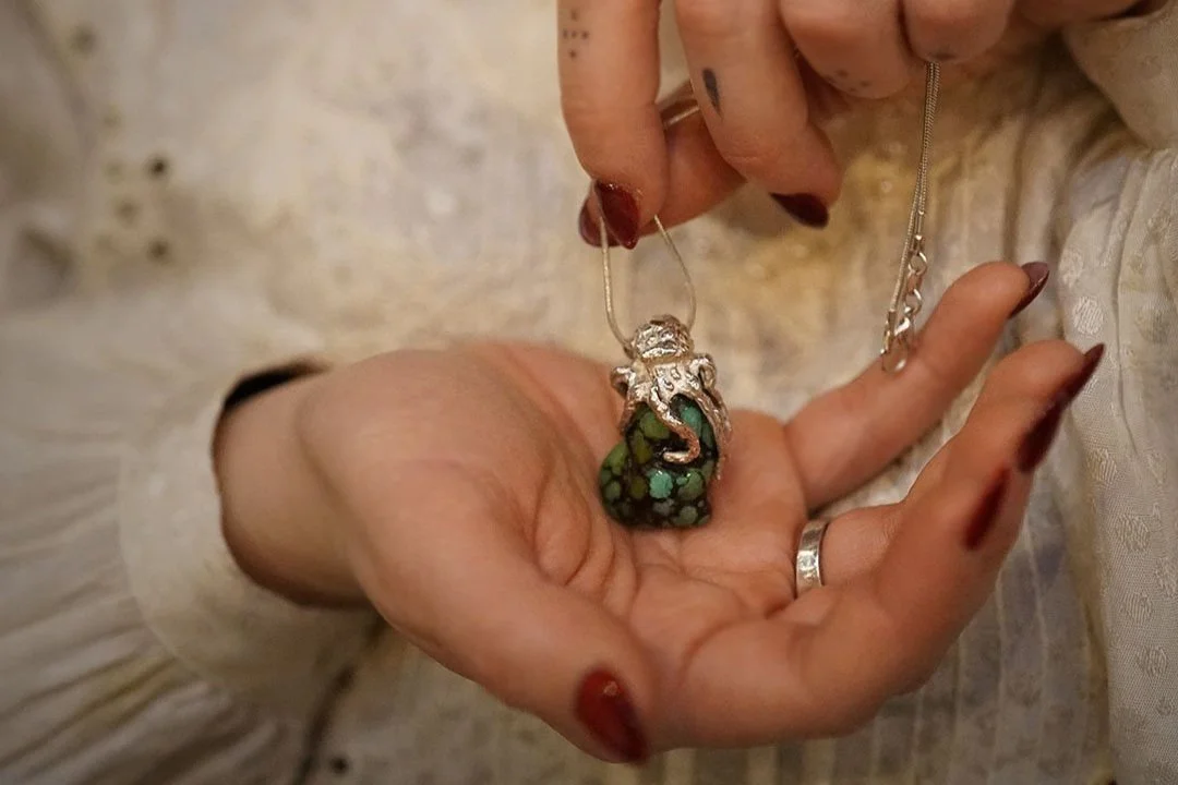 Main tenant un collier avec un pendentif en forme de pieuvre en argent, enroulé autour d'une turquoise. La personne porte aussi une bague en argent massif 925, avec des doigts manucurés en rouge.