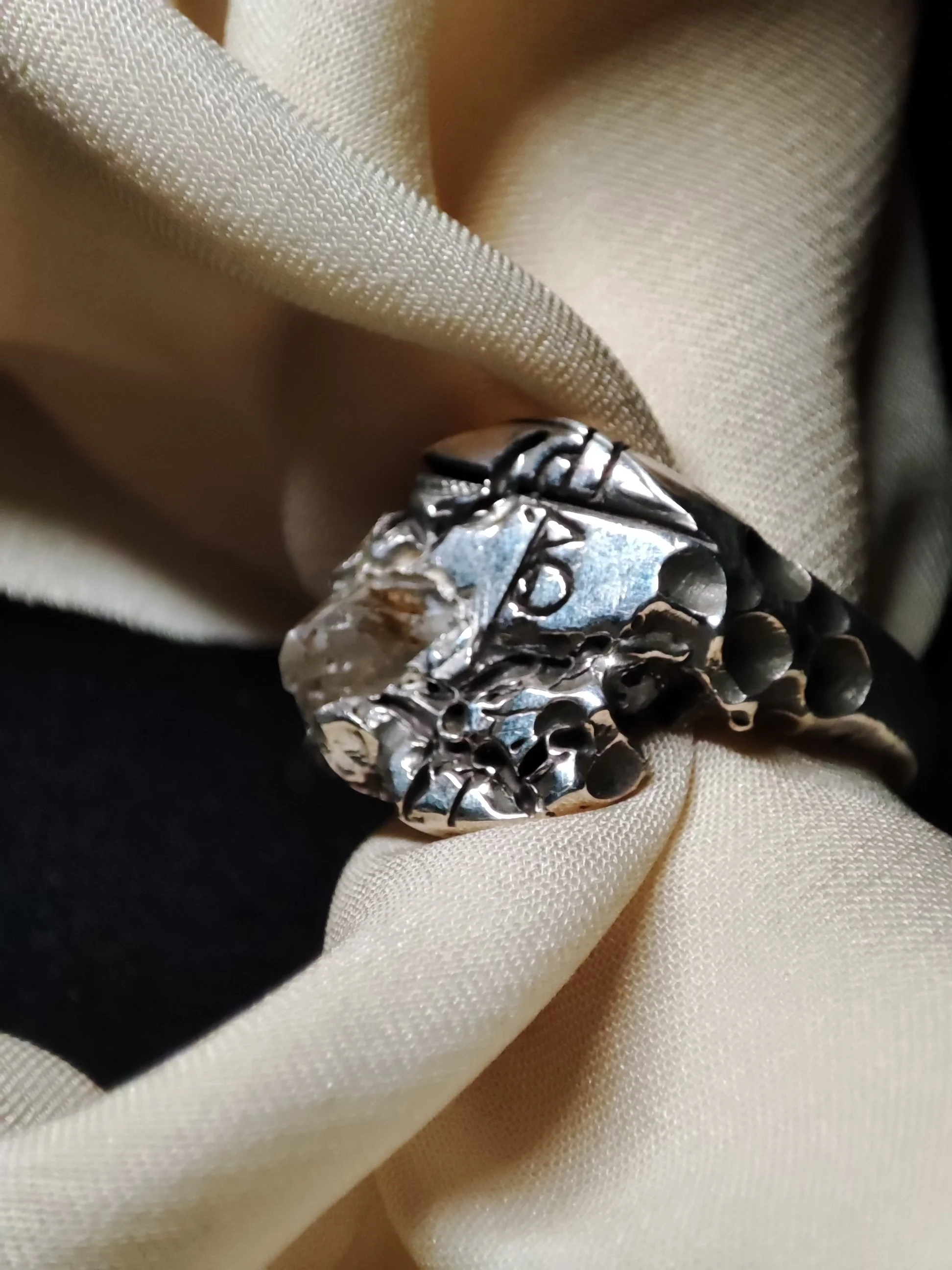 Une bague en argent massif 925 création originale de WIT joaillerie de la collection impacte avec un quartz pétrole, quartz à inclusion de pétrole naturelle du Pakistan réactif aux UV, posée sur un tissu beige.