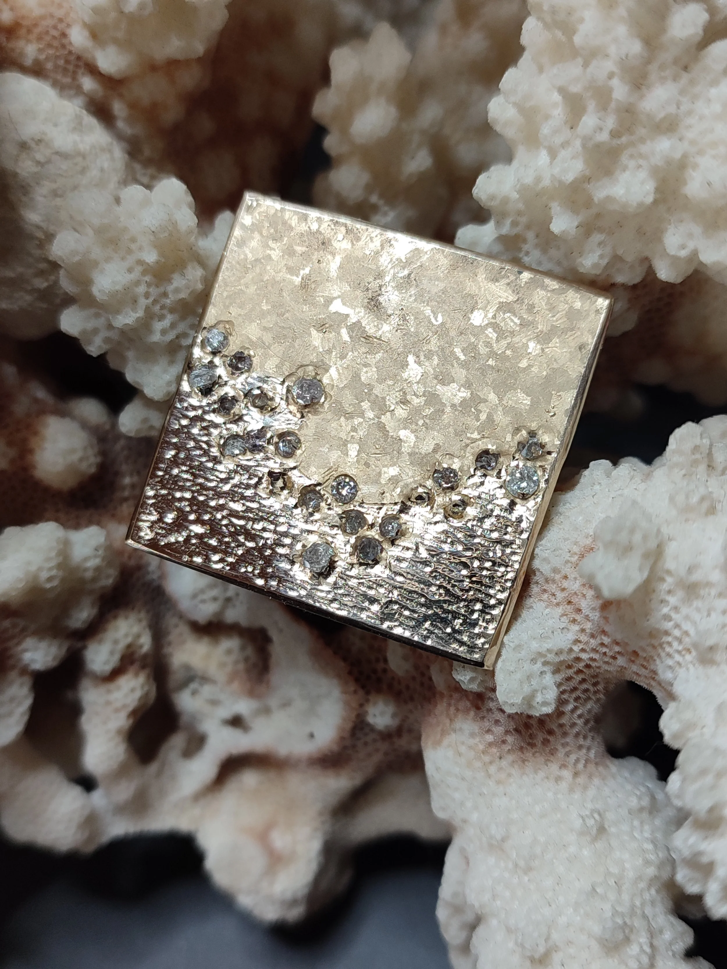 Un bijou en forme de carré avec des détails en relief et des pierres incrustées, posé sur du corail blanc.