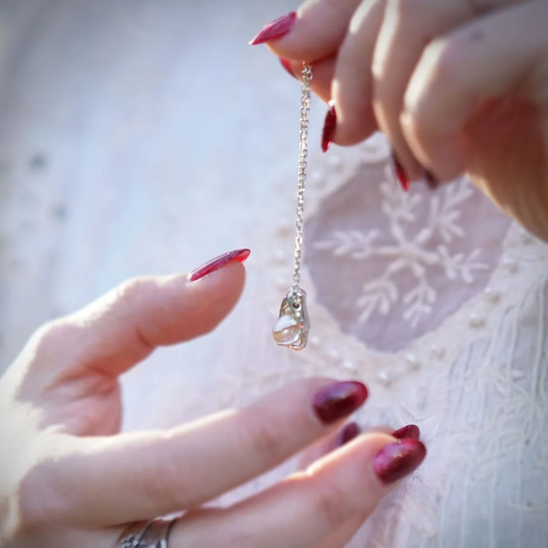 Mains féminines tenant une chaîne avec un pendentif en argent massif 925 en forme de main enserrant une perle, bijoux raffinés, fond flou. Fabrication française de bijoux de créateur.