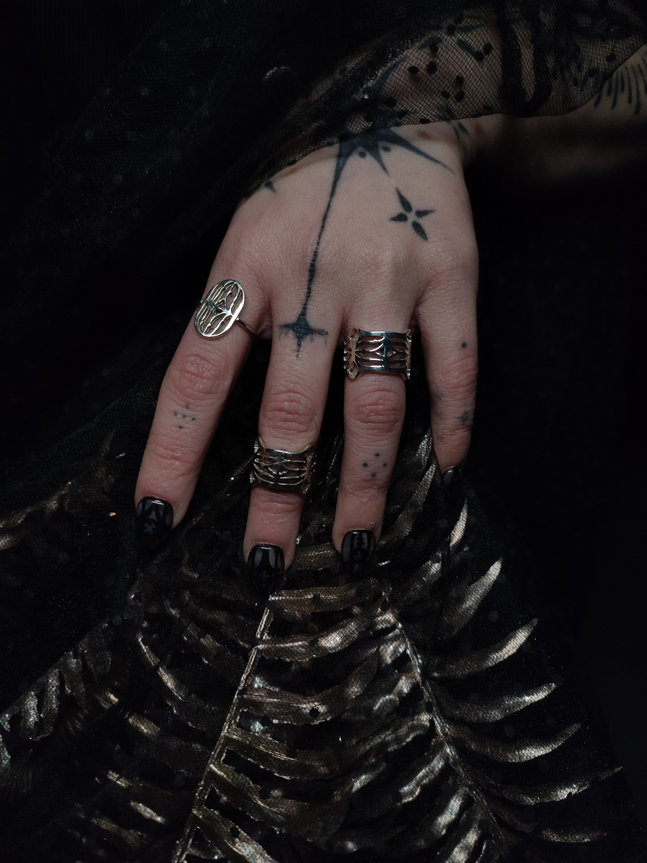 Main tatouée avec plusieurs bagues en argent massif 925, collection Cluny, motif s'inspirant du Musée de Cluny, Musée nationale du Moyen-âge de Paris. Fond sombre, ambiance gothique et végétale.


