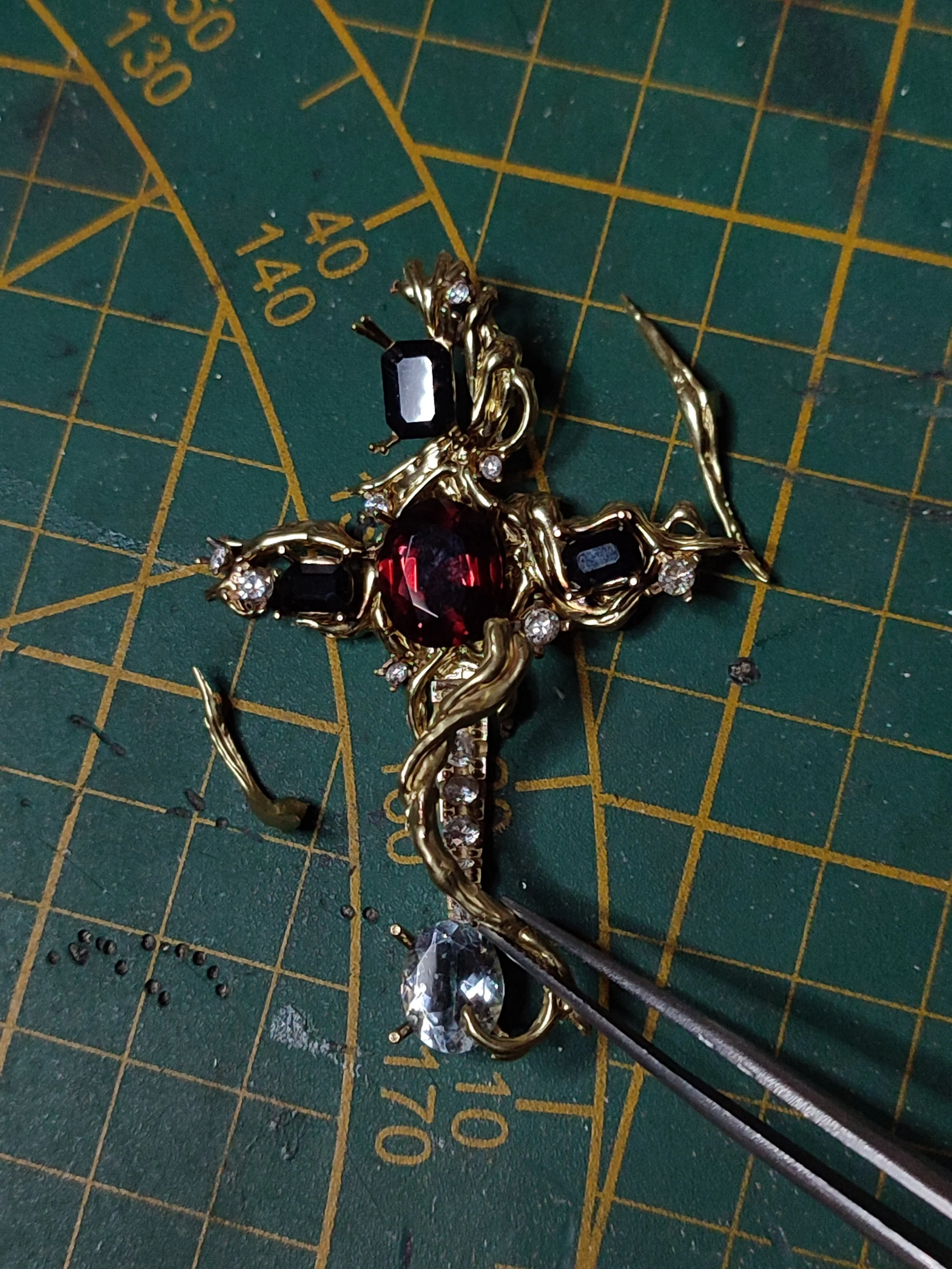 Pendentif ornementale en or en forme de croix avec pierres précieuses, posée sur un tapis de coupe avec des mesures.