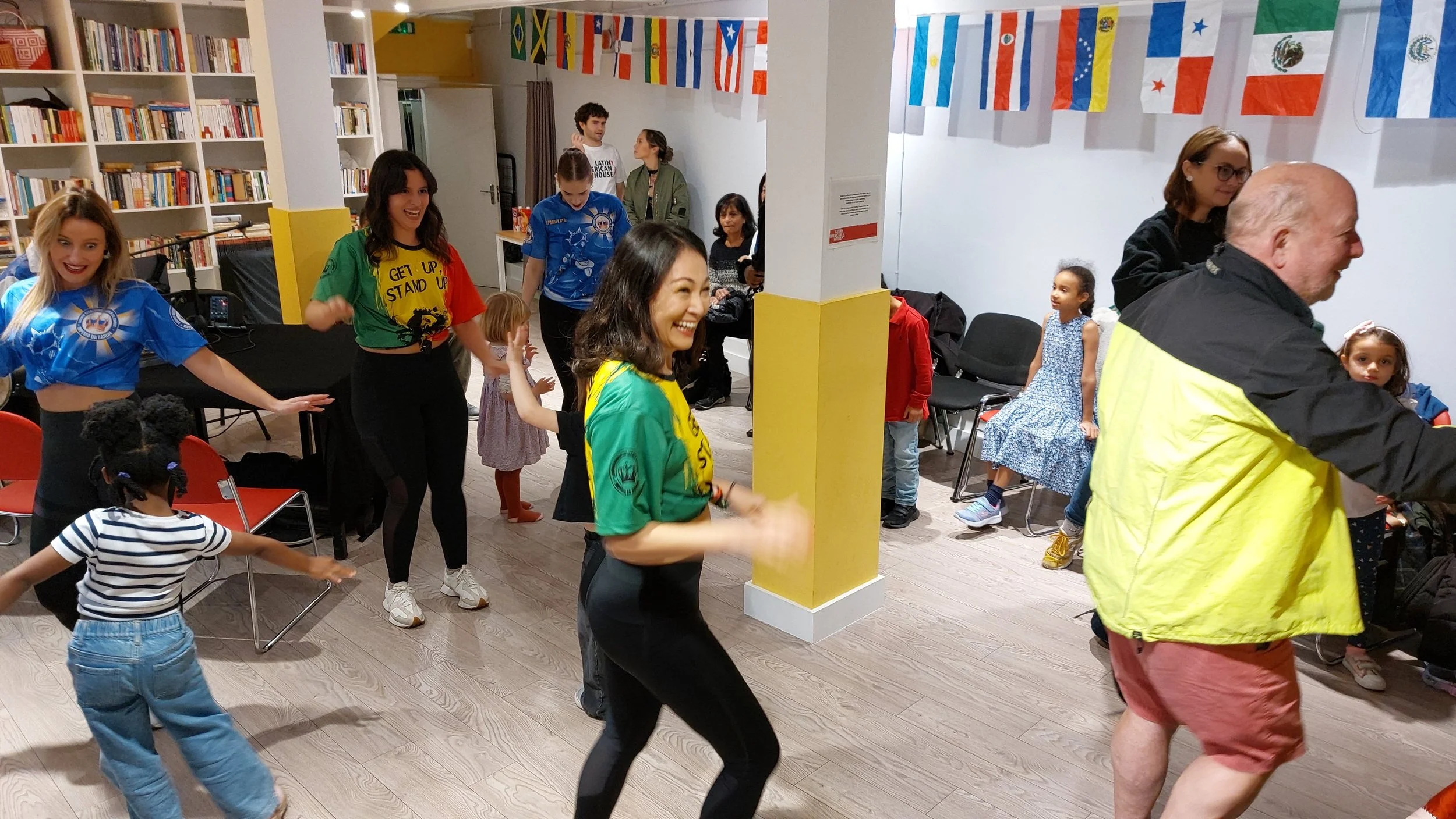 People dancing and enjoying themselves at an indoor event, with flags of various countries hanging on the wall and children and adults participating.