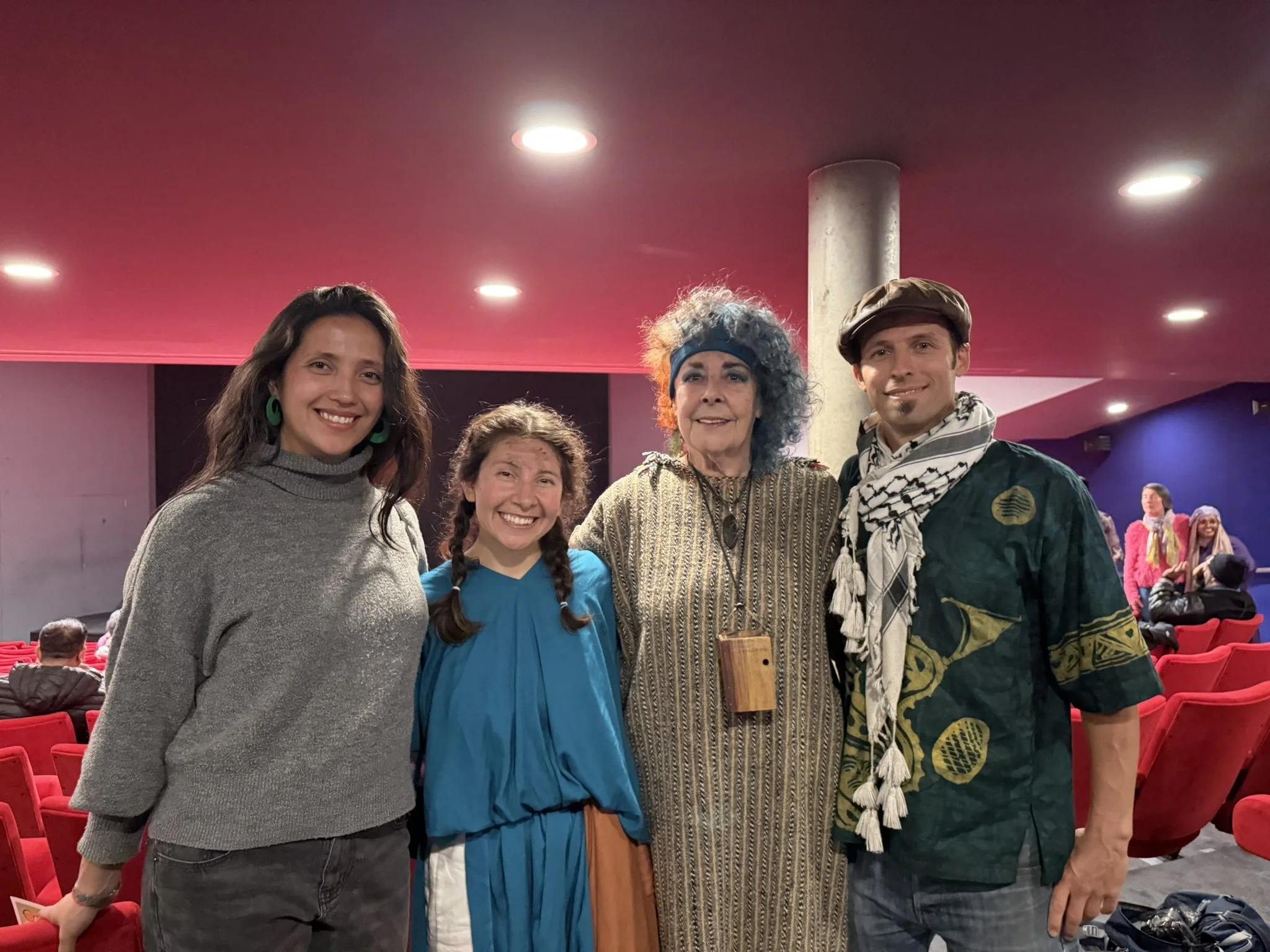 Four people smiling and posing for a photo in an indoor auditorium with red seating. The group includes two women with long hair, a woman with curly blue hair, and a man wearing a beret and scarf. In the background, other people are standing and sitt