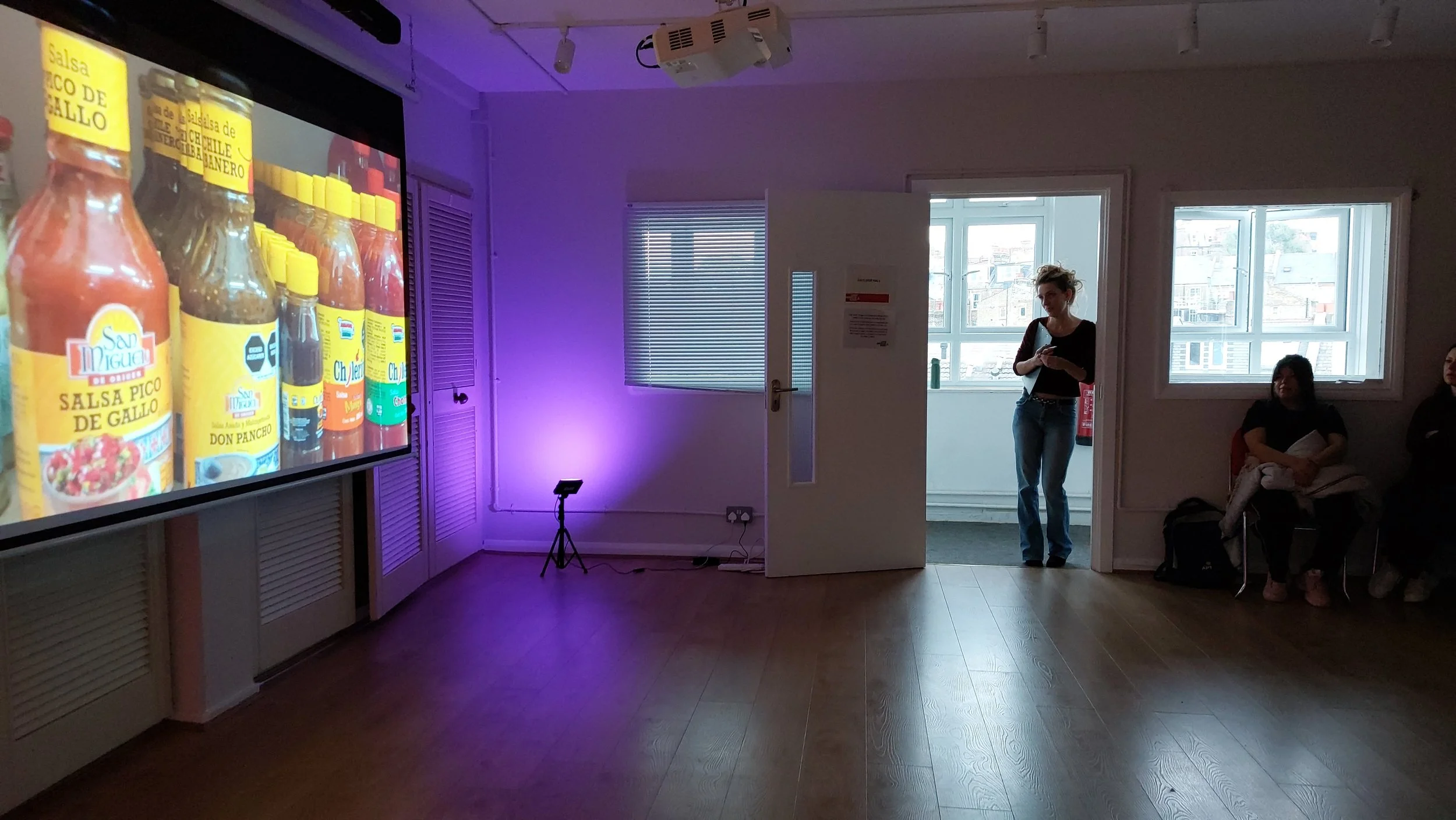 A room with a presentation screen on the left showing bottles of hot sauces. A woman stands near a white door, looking at her phone. Two women are sitting on chairs near the window on the right, one holding a notebook and the other resting with her a