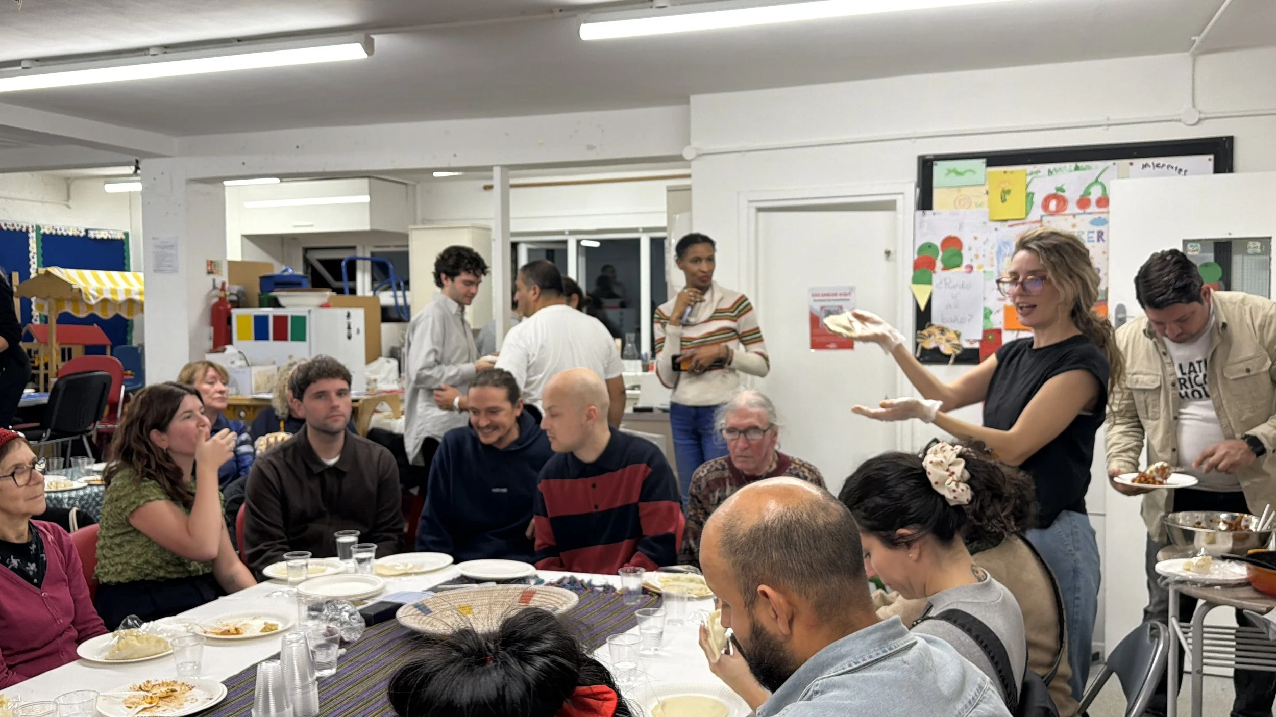 People gathered around a table at a social event, with some serving food and others eating, in a room with white walls and colorful decorations.