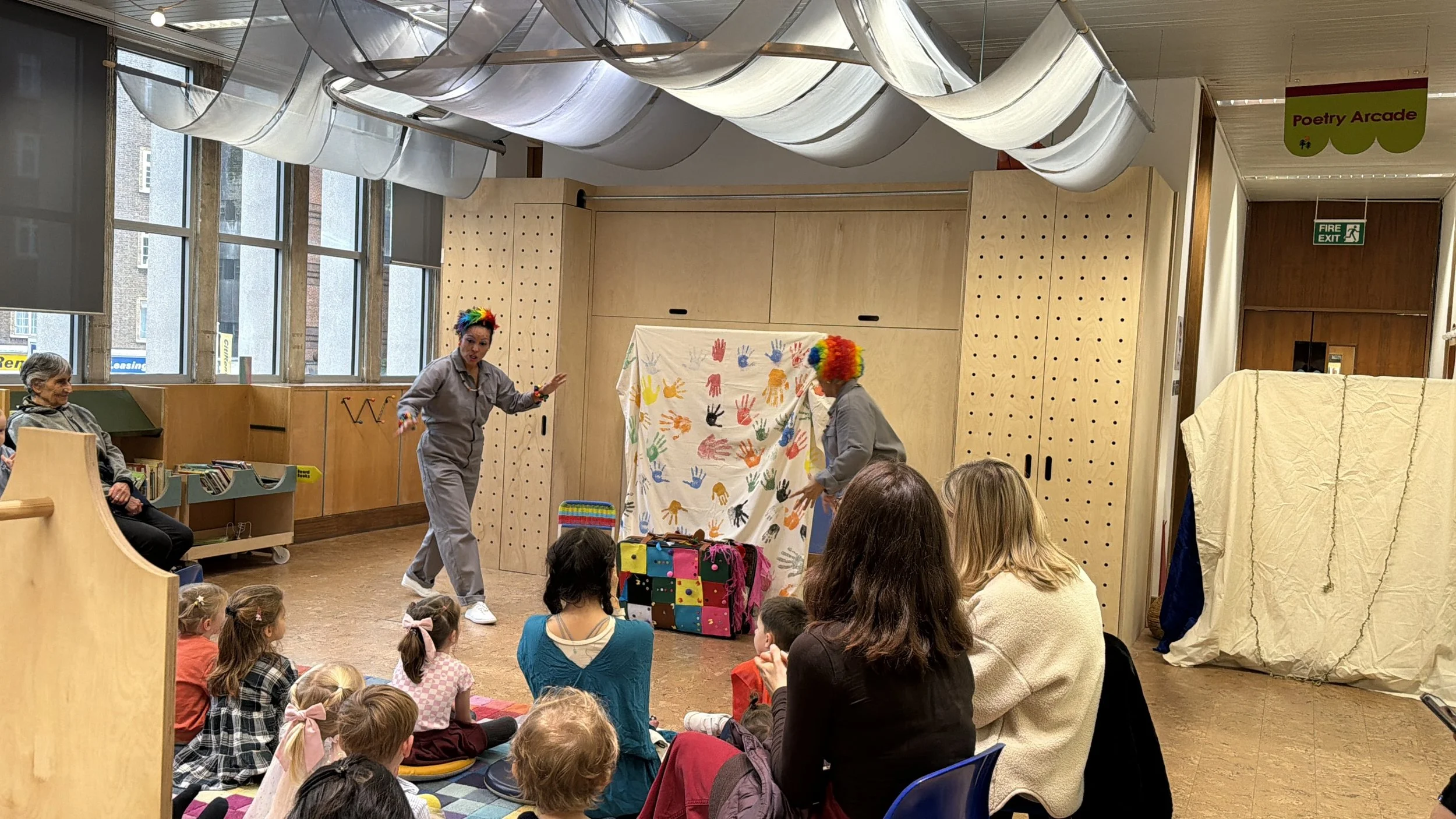 Children and adults watch a clown show in a library or community center, with two performers wearing rainbow wigs and gray jumpsuits, performing a puppet show using colorful handprints on a white sheet as a backdrop.