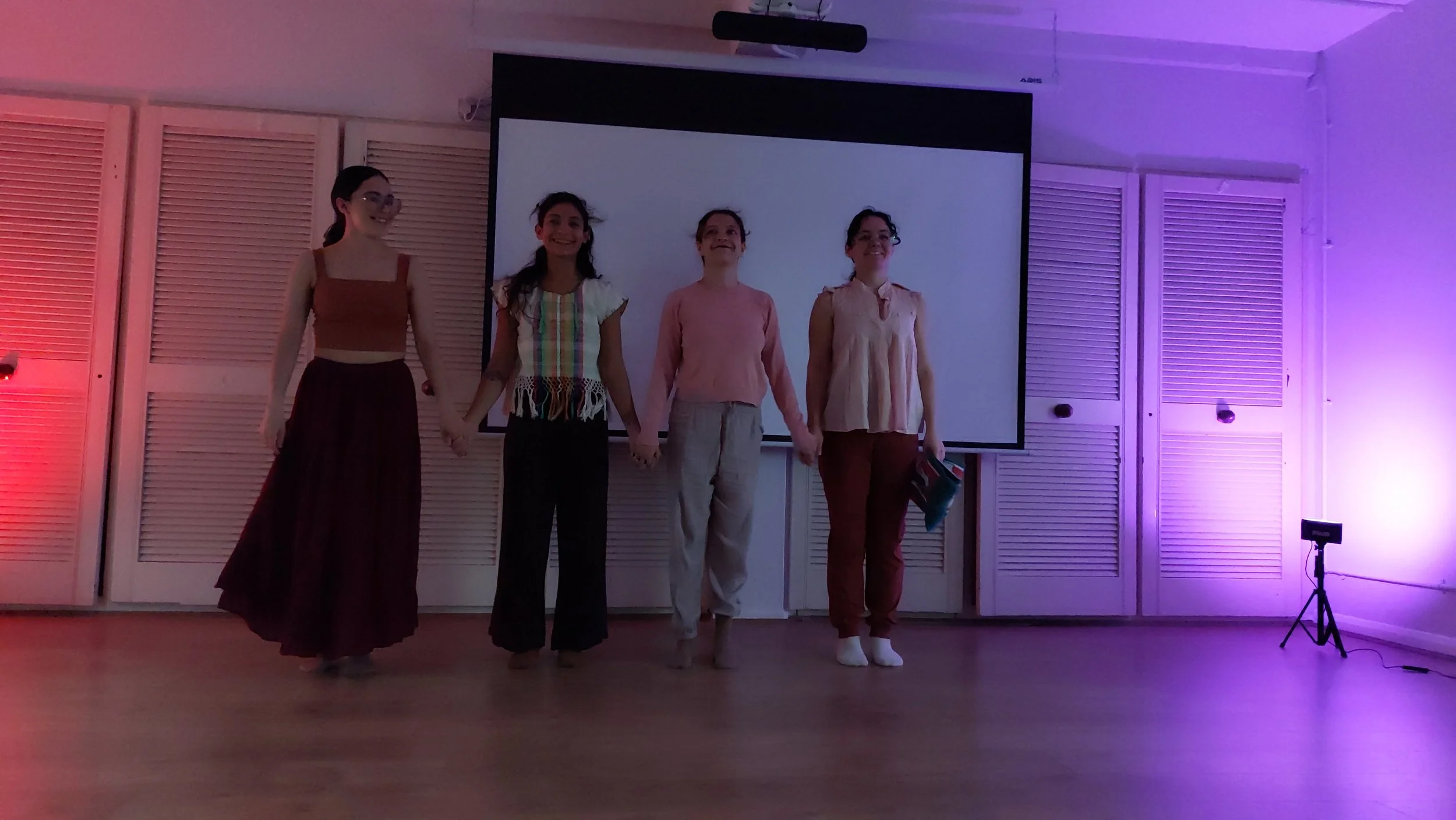 Four women are holding hands on stage in front of a large blank screen, with purple and red lighting illuminating the space. One woman is holding a folder, and they are smiling.