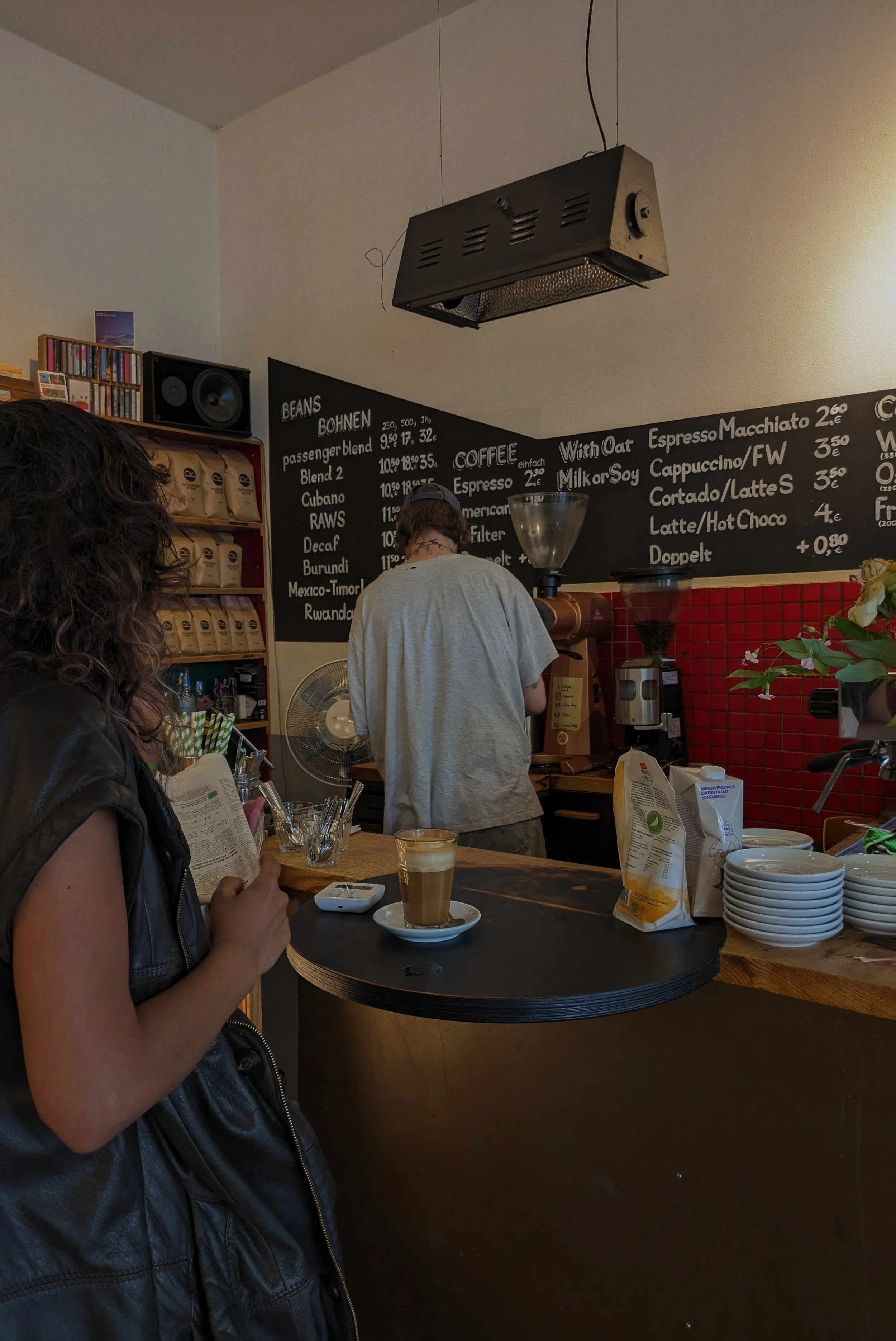 A woman standing at a coffee shop counter holding a menu, with a latte and smartphone on the counter. A barista is preparing drinks behind the counter, with a chalkboard menu in the background listing coffee options and prices.