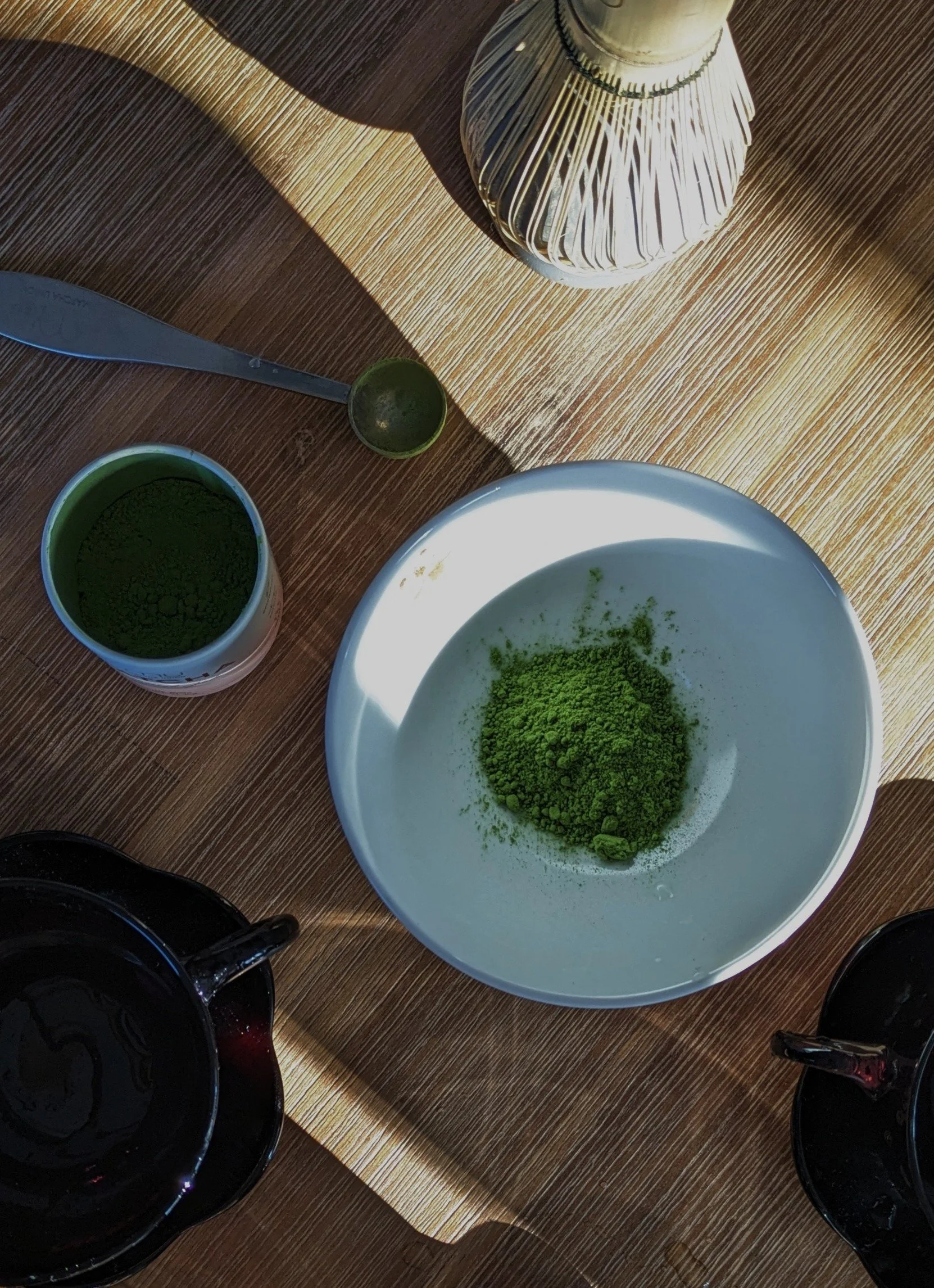 A wooden table with a white bowl containing green powder, a small green container, a jar of green powder, and two black cups, one filled with a dark liquid, possibly matcha tea.