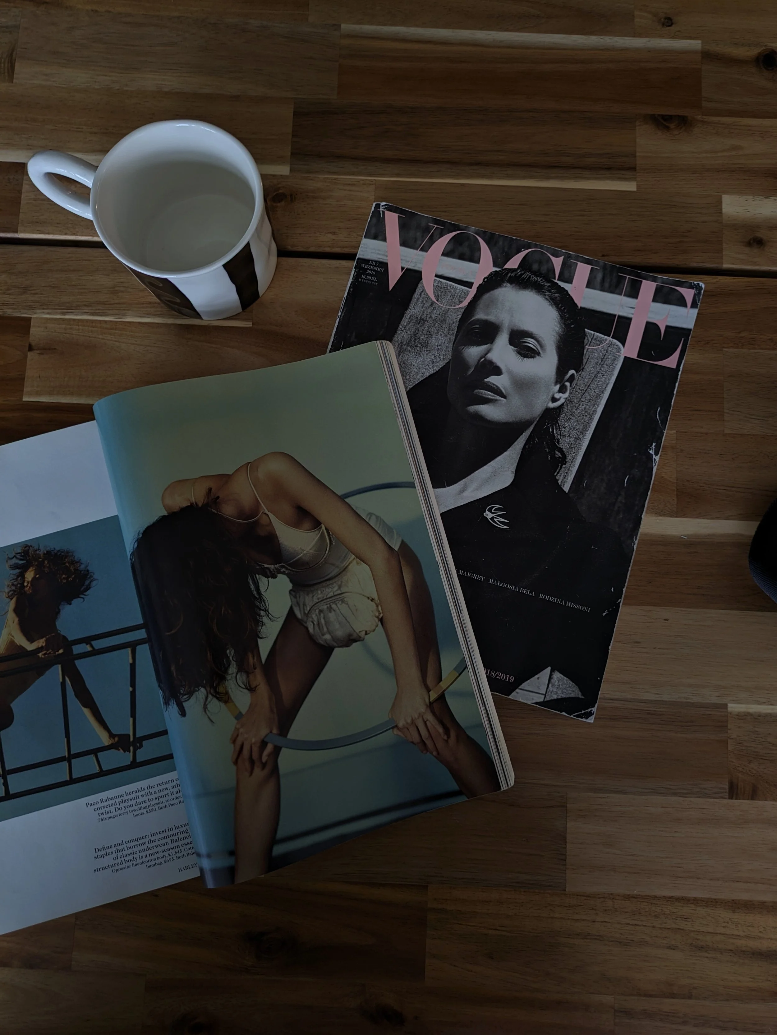 A wooden table with a white mug, a Vogue magazine with a black-and-white cover featuring a woman, and an open magazine showing a woman in a beige outfit posing with a hoop.